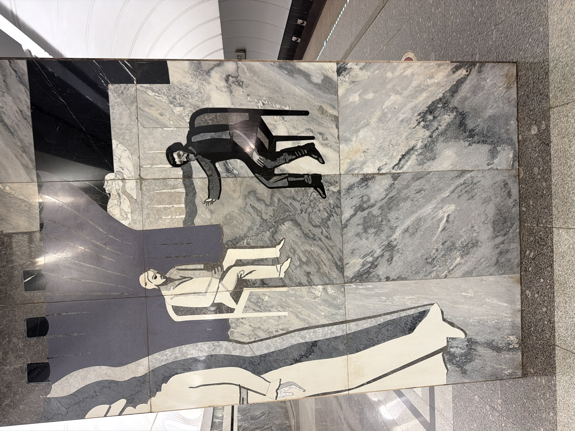 This image depicts a marble mosaic art installation on a pillar in the Moscow Metro. The mosaic features stylized figures of people rendered in contrasting light and dark stone. On the left, a tall, slender figure in a long garment with delicate hands is shown in profile, with flowing lines suggesting movement. To the right of this figure, two men are seated. The first man, on the left, wears a suit and hat and is shown with his head bowed, hands clasped in his lap. The second man, on the right, is depicted with a beard and is gesturing with his outstretched hand towards the first man. The background of the mosaic is a textured dark blue, with abstract grey shapes that create a sense of depth and shadow. The pillar itself is clad in marbled stone, with variations in color and pattern. In the background, the curved white ceiling and platform of a Moscow Metro station are visible, along with a portion of the track and signage. The overall impression is one of a public artwork in an underground transit environment.