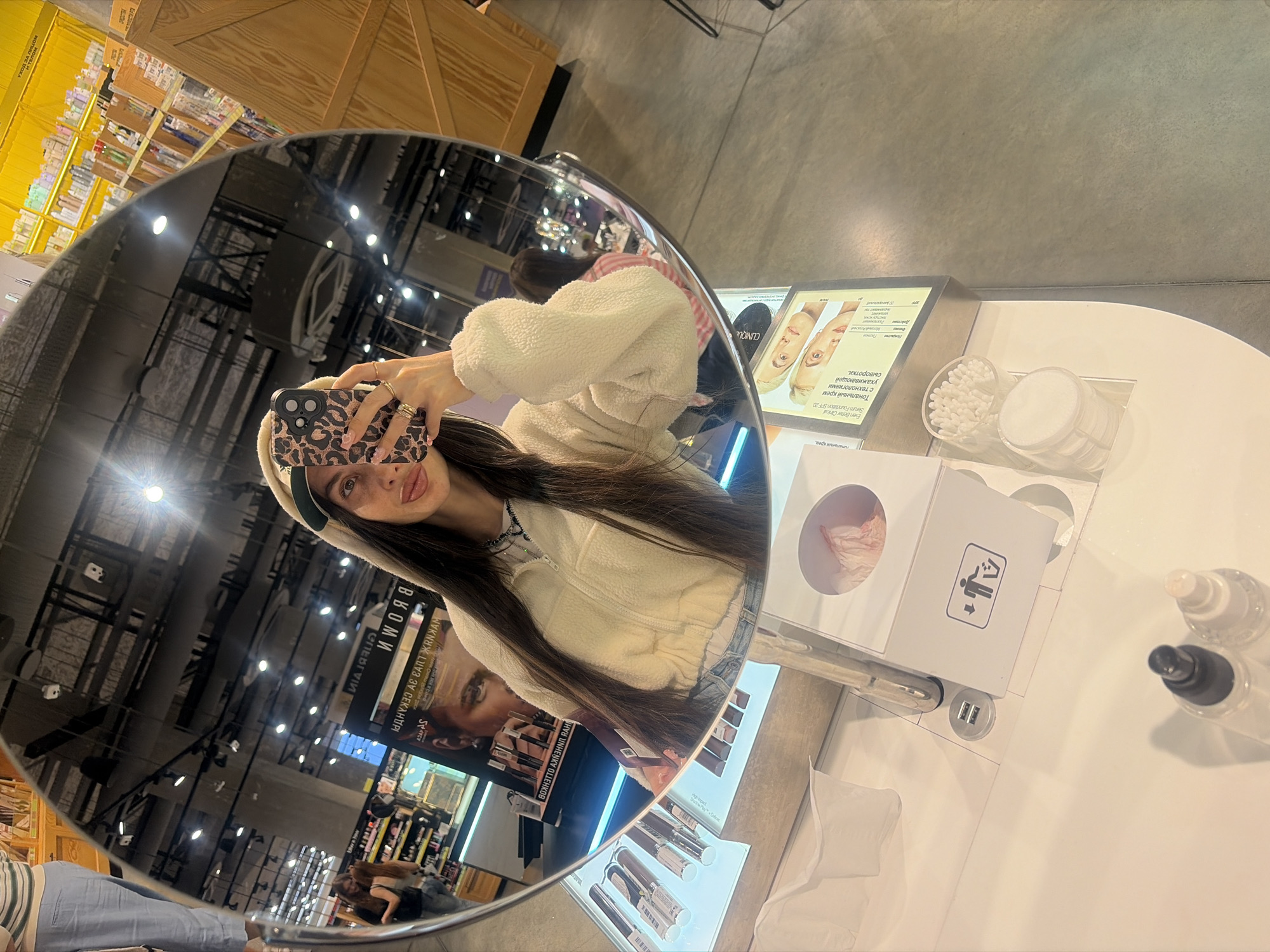 This is a medium-shot, reflective selfie taken in a store, likely a cosmetics or beauty shop, in Saint Petersburg, Russia. The main subject is a young woman with long dark hair, wearing a beige fuzzy jacket and a beige beret. She is holding a phone with a leopard-print case, angled to capture her reflection in a large, round mirror. The mirror distorts the reflection, showing a wider view of the store.
The store appears to be well-lit with bright overhead lights and displays of products. In the background, shelves are stocked with various items, and signage is visible. The woman's reflection shows her looking at the camera, smiling with her tongue slightly out, and wearing rings on her fingers. She has on light-colored jeans and a necklace.
In the foreground of the reflection, on a white counter, are several beauty products. These include cotton pads, cotton swabs, and bottles of skincare or makeup. A digital display shows images of faces and text in Russian, possibly advertising a "Clinique" product. The Russian text on the display reads "Тональный крем с технологиями ухаживающей сыворотки" (Foundation with caring serum technologies) and "Even Better Clinical Serum Foundation SPF 20". Another sign in the reflection reads "ВКОМИ".
The overall mood is casual and playful, with the woman seemingly enjoying herself while taking the photo in the store environment. The lighting suggests it's daytime, or at least indoors with strong artificial lighting. The image is rich with detail due to the reflections in the mirror.