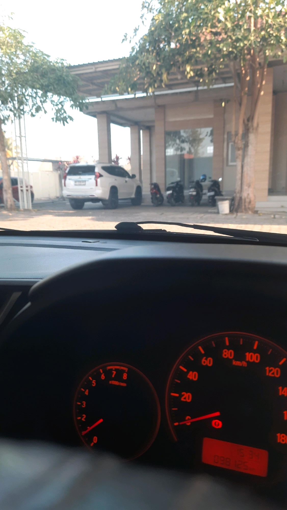 In the city of Jajag, Indonesia, a view from inside a car captures a scene outside through the windshield. A white SUV is parked in front of a building with a covered outdoor area supported by pillars. Several motorcycles are also parked near the building. Trees with sparse foliage are visible in the foreground and around the building. The car's dashboard, with illuminated gauges and a digital display, is in the foreground, indicating speed and other vehicle information. The overall lighting suggests it is daytime, with bright sunlight casting shadows.