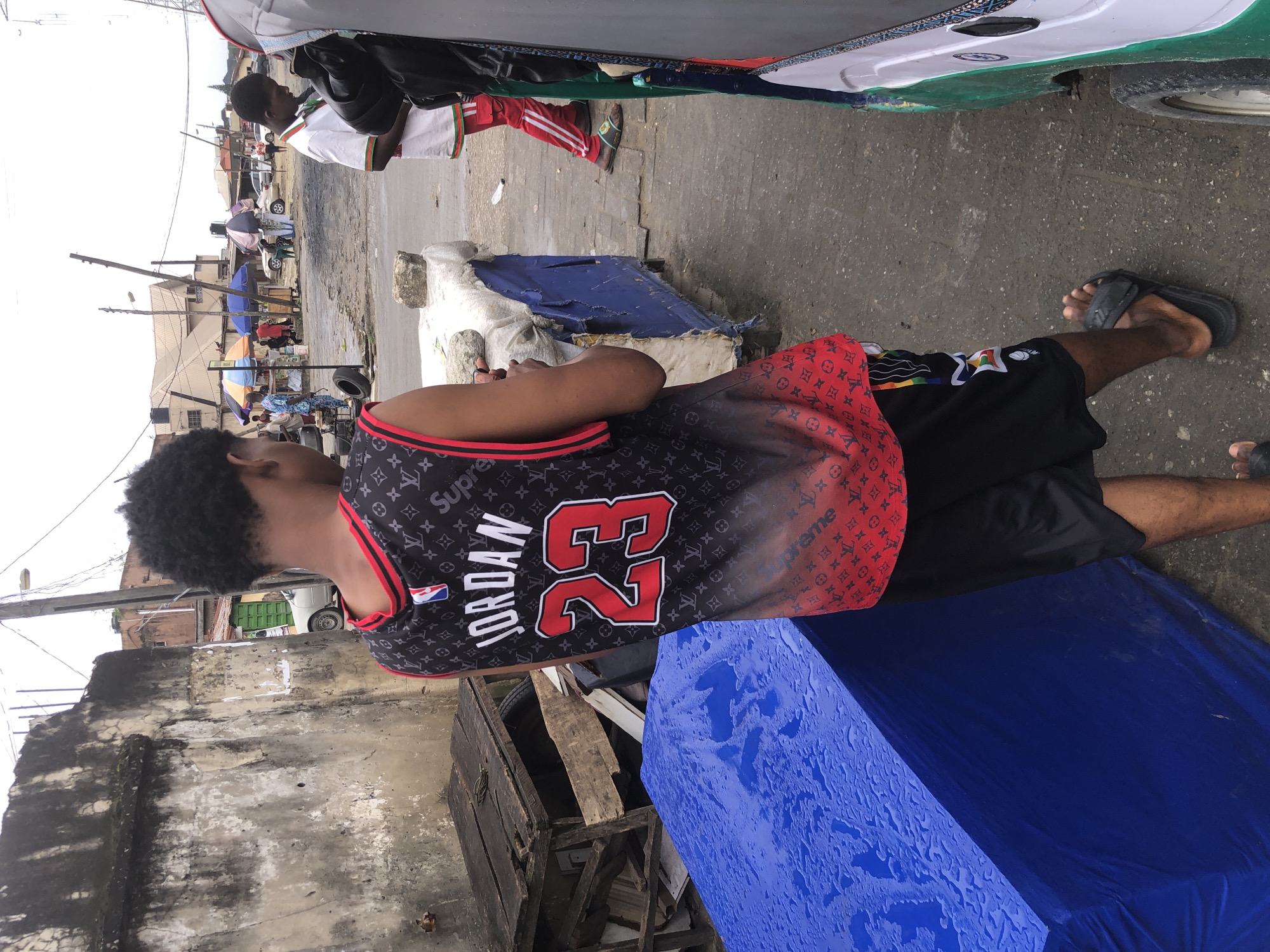 Ado, Nigeria. The image captures a street scene with a young Black male in the foreground, facing away from the camera, wearing a black basketball jersey with red trim and a "Jordan 23" graphic, along with a Louis Vuitton-inspired pattern. He is also wearing black shorts with rainbow-colored accents and black sandals. To his left, a large blue tarp covers an object, and behind him, a weathered concrete wall stands. In the background, market stalls with colorful umbrellas are visible, along with people, vehicles including a tuk-tuk, and buildings. The ground appears to be wet, suggesting recent rain.