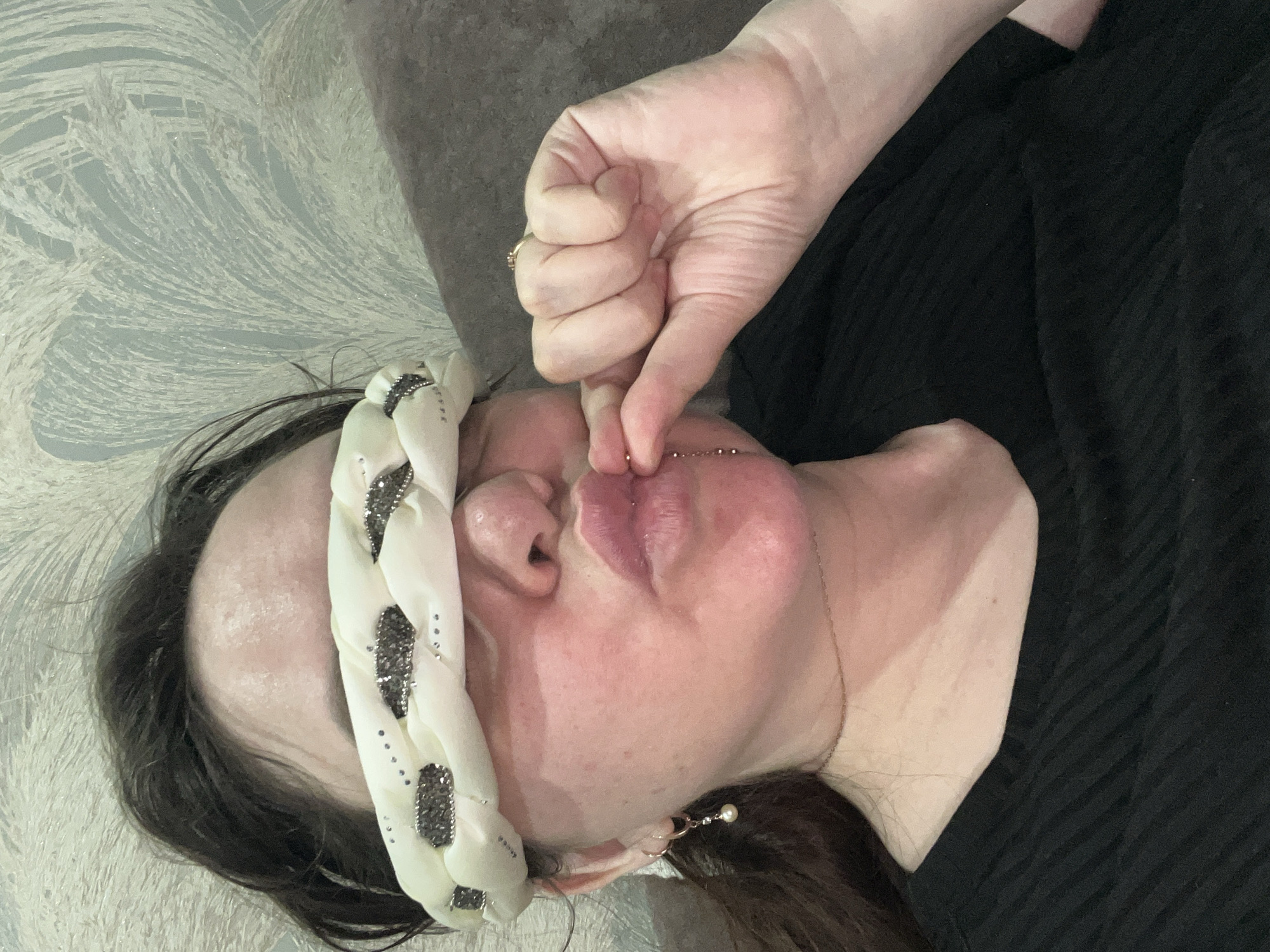 A supine adult female with light skin is positioned against a light blue patterned surface. She wears a white braided headband decorated with dark, square-shaped embellishments. Her dark hair is visible at the lower right of the image. She wears earrings. An adult hand with pale skin, partially obscuring the subject’s face, is pinching a chain near her lip, held between the thumb and index finger. The background includes a gray-toned surface. The subject is wearing a black top. The scene's location within Noviy, Russia is unknown.