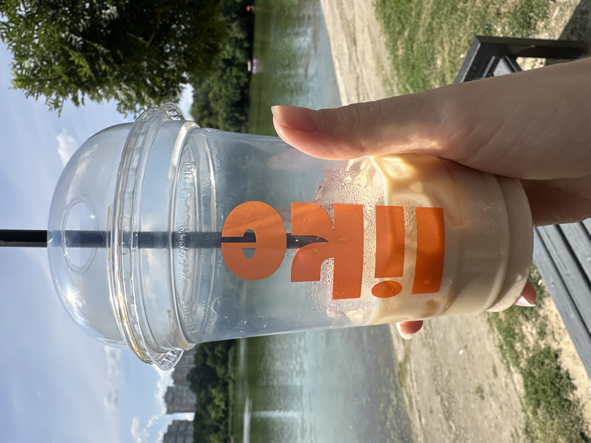 A hand holds a clear plastic cup with an orange logo and a straw, containing a light-colored beverage. The cup is covered in condensation. Behind the cup, a body of water with trees lining its edge and some buildings in the distance is visible. The sky is blue with some clouds. A wooden bench and a grassy area are also present in the background, suggesting an outdoor setting. The overall impression is that of a refreshing drink enjoyed on a pleasant day, likely during daylight hours. The orange logo on the cup appears to spell out "I LIKE" or a similar phrase, with an exclamation point. The image has been taken outdoors, in what appears to be a park or a similar recreational area, possibly near a lake or river. The weather seems to be sunny and warm, given the condensation on the cup and the clear sky. The presence of buildings in the distance and the lush greenery of the trees suggests an urban or suburban environment. The focus is primarily on the drink and the hand holding it, with the background providing context to the setting. The overall mood conveyed by the image is one of relaxation and enjoyment.