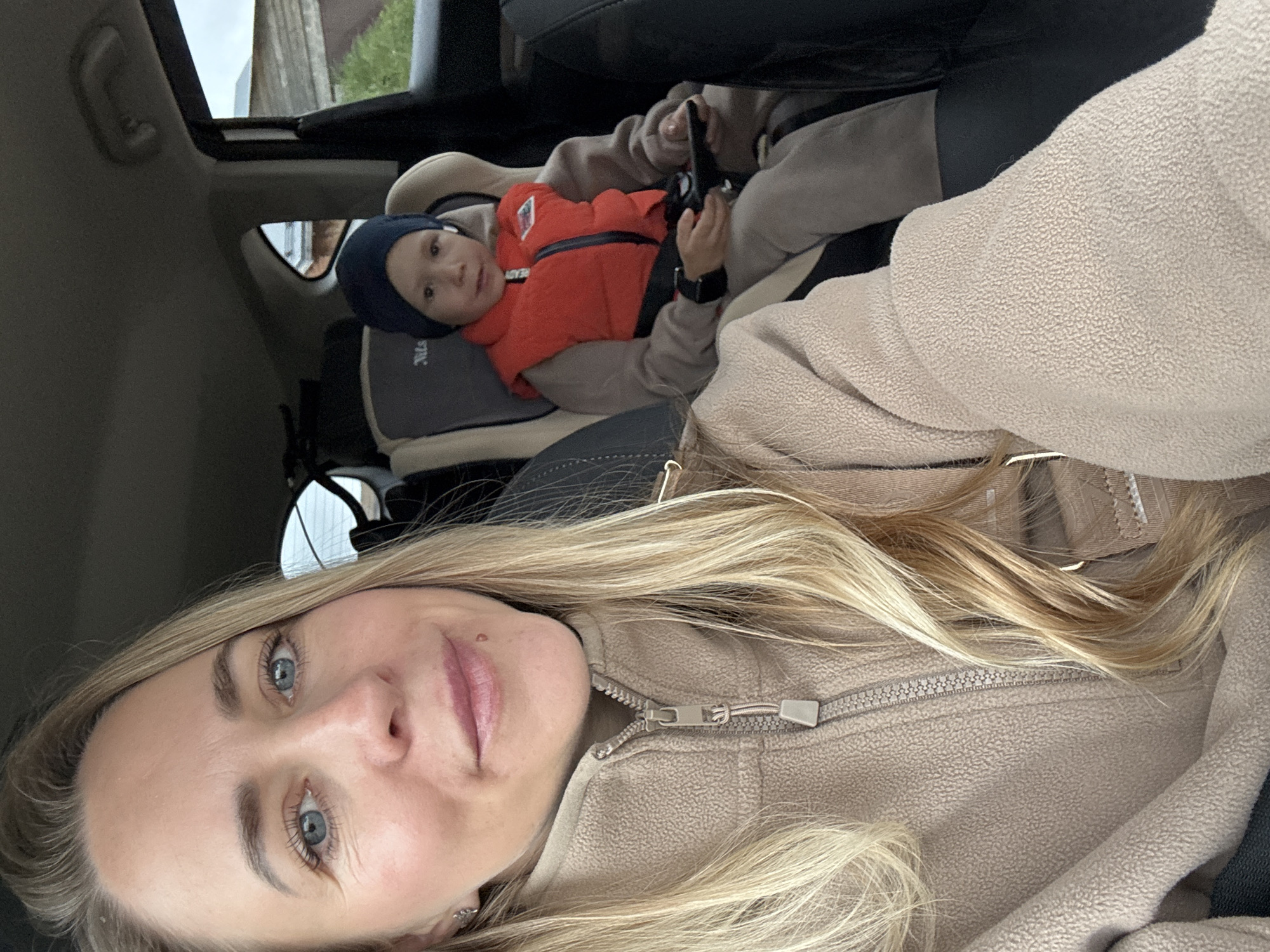 A woman with long, blonde hair wearing a tan fleece jacket is taking a selfie in the back of a car. A young child wearing a navy blue hat and an orange vest is strapped into a gray car seat behind her, looking at the camera. The car has a sunroof. The image was likely taken in the city of Chaikovsky, Russia based on the request.