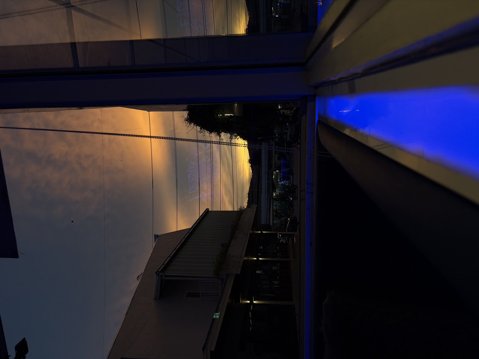 A dimly lit outdoor scene at dusk, viewed through a window. The sky is a deep blue transitioning to orange clouds at the horizon. A modern building with a light-colored facade and dark metal siding is visible on the left, featuring large windows and a covered walkway with illuminated lights. A string of beads hangs vertically in the foreground, reflecting the sky. In the middle ground, there are streetlights and what appears to be a road with several motorbikes parked or moving. A bright blue light emanates from a strip along the bottom of the window frame. The reflection in the window shows the orange sky, trees, and more streetlights.