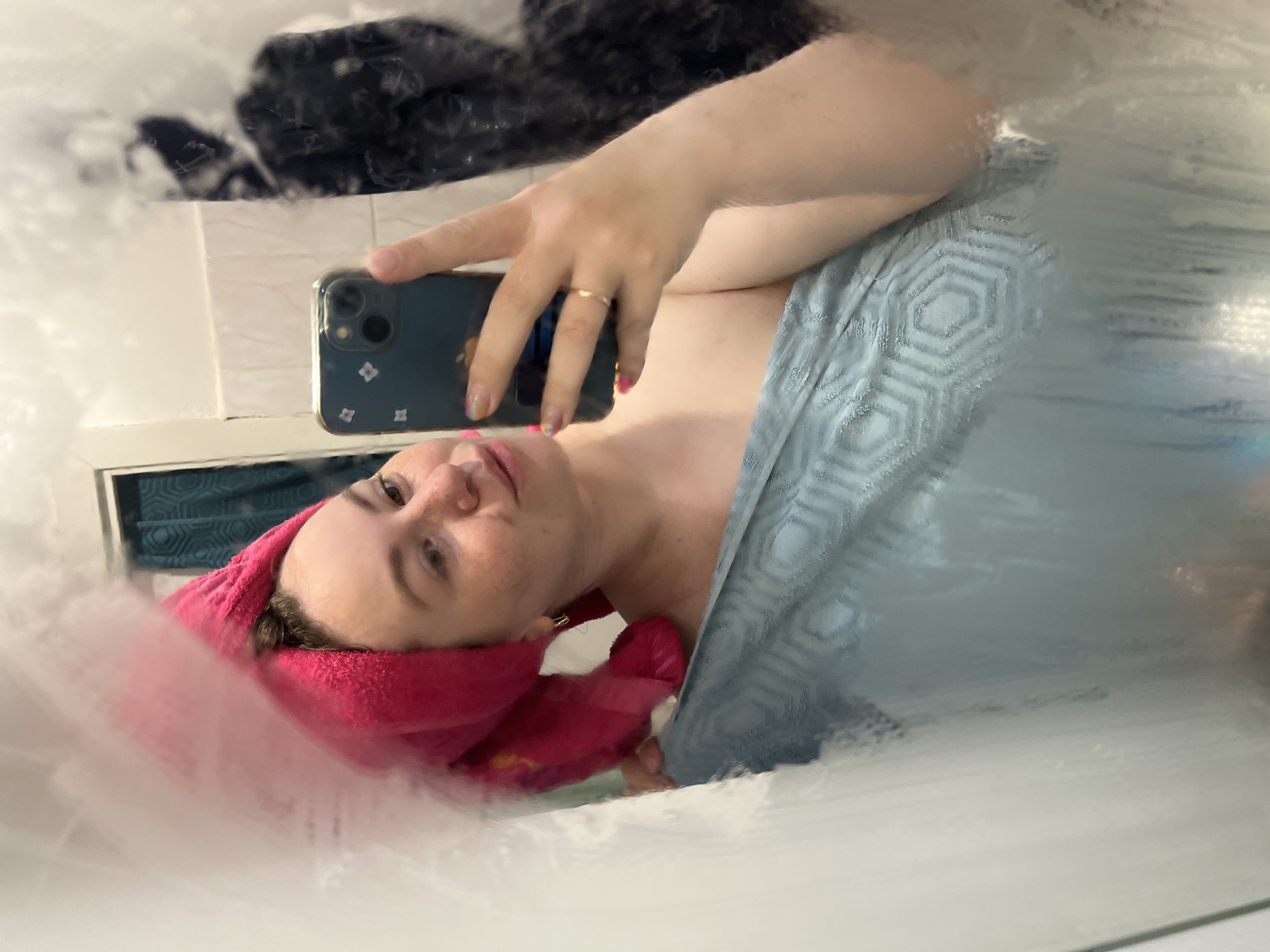 A mirror selfie shows a woman wearing a pink towel on her head and a blue robe. She is holding a dark blue iPhone with a floral design on the back in her right hand, taking the picture. She wears a gold ring on her right ring finger. The mirror is steamy. Behind her, in the mirror's reflection, is a white tiled wall and a black object. The location is likely a bathroom. There is no information to confirm this image was taken in Chaikovsky, Russia.