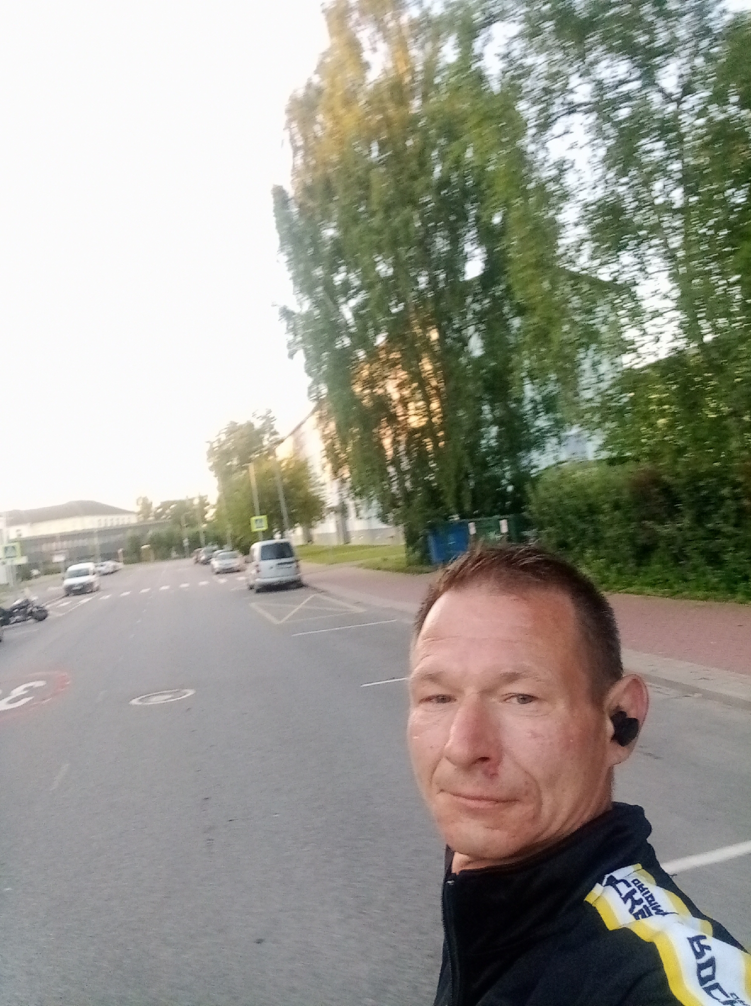 A man is taking a selfie while standing on a street in Tapa, Estonia. The man is wearing a black jacket with yellow and white stripes on the sleeve and black wireless earbuds. He has short brown hair and is looking at the camera. In the background, there is a street with several cars, a sidewalk, and trees. There is also a building in the distance. The sky is overcast.