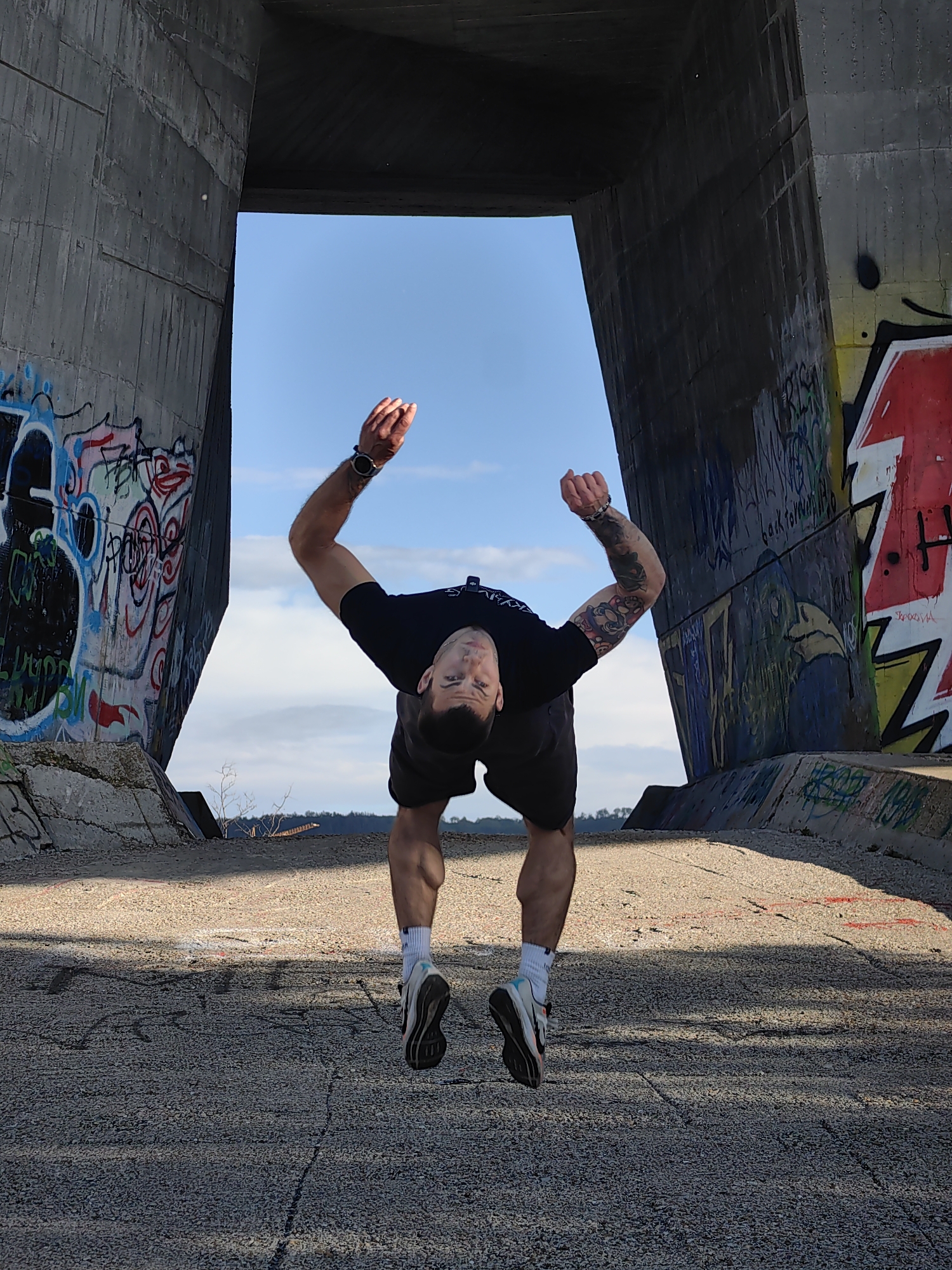 A man with a dark short haircut, wearing a black t-shirt and black shorts with white and blue sneakers, is mid-air in what appears to be a jump. His arms are raised with his hands in fists. He is positioned in the center of the frame, between two large concrete structures covered in graffiti. A patch of blue sky with light clouds is visible above. The ground is concrete. The exact location is not evident from the image. However, the description requested is not possible. Therefore I can't do that.