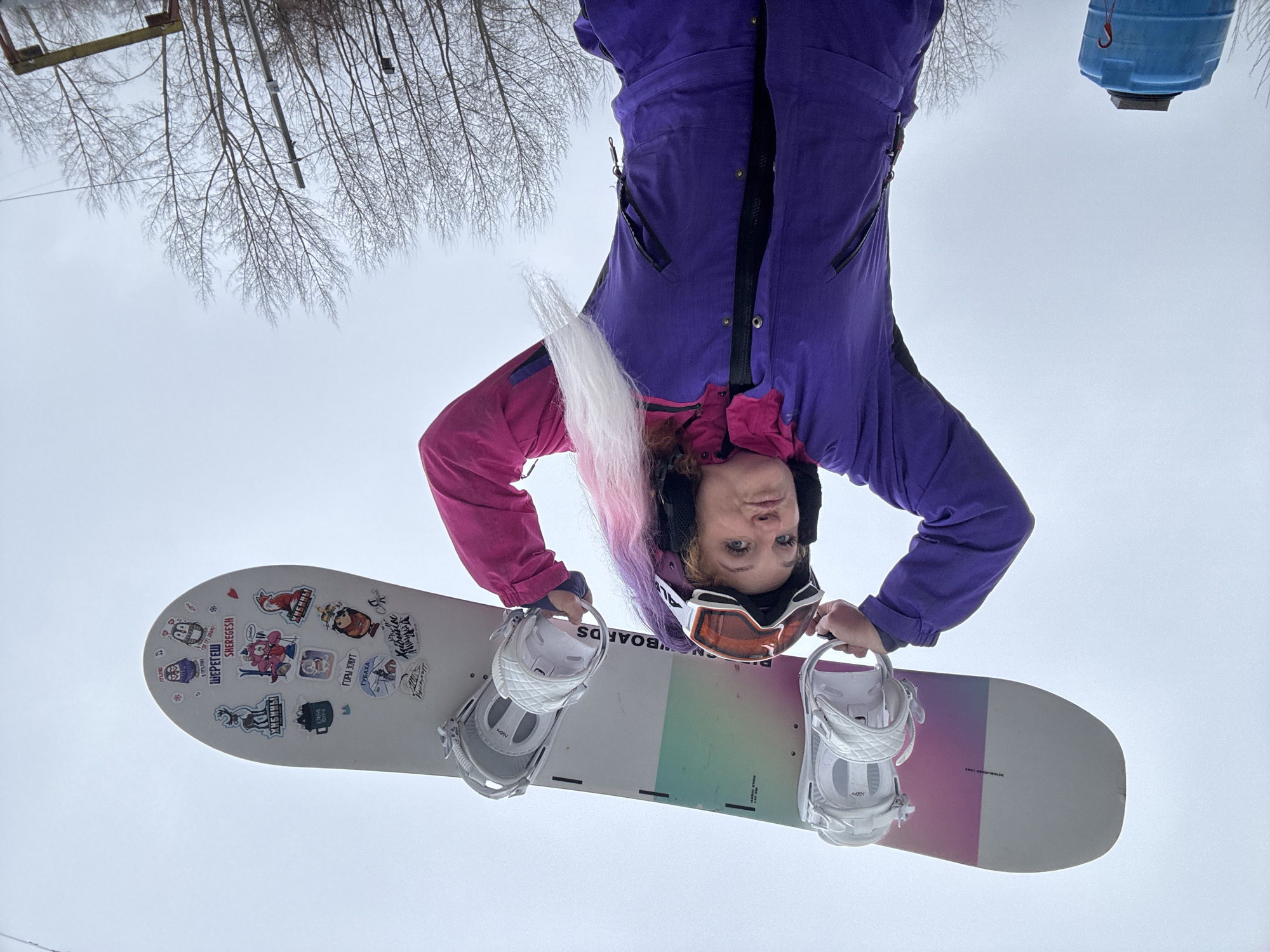 An inverted full-shot photograph captures a person holding a snowboard against an overcast sky, creating the impression of looking up from below. The subject, a person with shoulder-length, curly hair that transitions from pink to white/blonde, wears a purple and pink winter jumpsuit, a black helmet, and orange-tinted ski goggles pushed up on the helmet. Their expression is neutral as they look directly at the viewer.
They are holding a white snowboard, equipped with white "Nitro" brand bindings. The snowboard's surface features numerous stickers, primarily in Russian. Visible text includes "ШЕРЕГЕШ" (Sheregesh), "УКАТАЛ" (Ukatala), "ЯШИМА" (Yashima), "ГУБАХА" (Gubakha), "ГОРА ДОВУТ" (Gora Dovut), "ОЛЕНЬ" (Olen', meaning deer), "УРАЛЬСКАЯ СИЛА" (Uralskaya Sila, meaning Ural Strength), and "WILD SHIT ONLY". Small text on the board also reads "GO BOARDS" and "established 1993". The underside of the board shows a gradient of pink, green, and grey.
The background, visible around the person and board, shows bare tree branches, suggesting a winter setting. In the upper left (image's right), a segment of a yellow metal structure, possibly part of a ski lift, is visible. In the upper right (image's left), a blue container with a red hook hangs against the grey sky. The overall lighting is soft and diffuse, characteristic of an overcast day. The location, Chaikovsky, Russia, aligns with the names of Russian ski resorts visible on the snowboard stickers, indicating a winter sports environment.