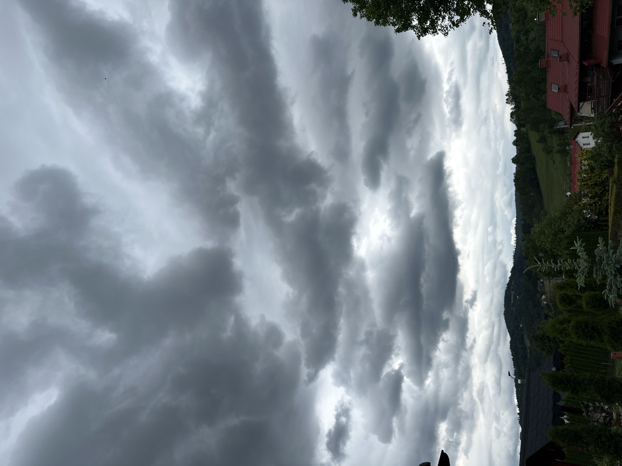 This image captures a dramatic and heavily clouded sky over a mountainous landscape in Wisła, Poland. The sky is the dominant feature, filled with layers of dark and light grey clouds exhibiting distinct, wave-like formations, possibly altocumulus undulatus. Brighter patches in the clouds indicate moments where sunlight is attempting to break through, creating high contrast. A tiny, dark bird is visible as a silhouette high in the sky in the upper left area.
Below the expansive sky, a verdant and inhabited landscape unfolds. On the right side of the frame, several houses or buildings with traditional reddish-brown tiled roofs and light-colored walls are nestled amidst lush greenery. Surrounding these structures are various trees and neatly trimmed bushes, suggesting gardens or cultivated areas. Further into the background, rolling hills and mountains, densely covered in dark green forests, stretch into the distance under the overcast conditions.
The scene conveys an atmosphere of unsettled or impending weather, likely a heavily cloudy or pre-storm day, though no rain is visible. The time of day appears to be daytime, with diffused light rather than direct sunshine. No specific activity is taking place, and there are no visible people or text. The image highlights the natural beauty of the Polish landscape under a powerful, dynamic sky.