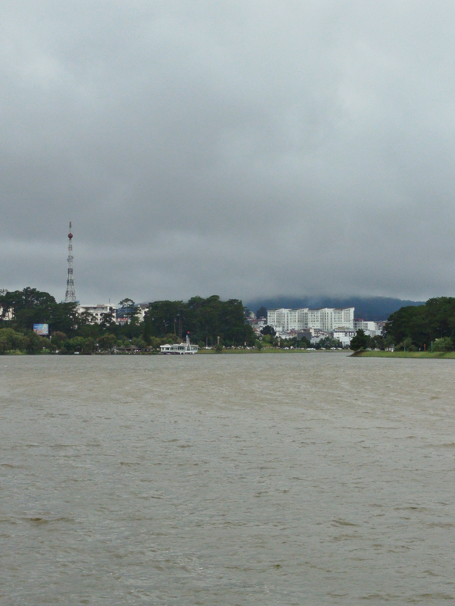 This image depicts Xuan Huong Lake in Da Lat, Vietnam, under an overcast sky. The lake occupies the foreground, its surface rippled by a gentle breeze. In the middle ground, a tree-lined shore separates the lake from a city skyline. A prominent radio tower, tall and lattice-worked with a red and white antenna at its apex, stands to the left of the frame. Behind the tower, a cluster of white buildings is visible. Further to the right, a long, multi-story white building complex dominates the landscape, with a row of smaller structures in front of it. A white, boat-like structure with a red-roofed cabin is moored along the shore, its white railings casting a shadow on the water. The distant hills are shrouded in mist, adding to the subdued atmosphere of the scene. The overall impression is one of a tranquil, slightly melancholic urban lakeside environment.
