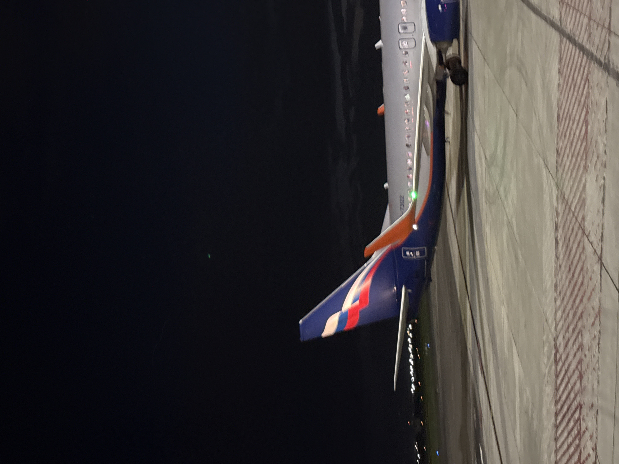 The image captures a night scene at an airport, featuring the rear and side of a large airplane oriented vertically along the right side of the frame. The left portion of the image is dominated by a dark, starless night sky, with a faint green light visible towards the upper left. The airplane's fuselage is primarily white, with a distinctive blue, red, and white livery on its vertical stabilizer (tail fin), strongly suggesting an Aeroflot aircraft. Part of the aircraft's registration number, visible on the fuselage below the wing, reads "-7322".
Brightly lit cabin windows are visible along the fuselage, and a prominent green navigation light is active towards the front of the visible section. The ground, likely a tarmac or taxiway, is visible on the far right, showing concrete paving with red and white striped markings. Faint distant lights suggest other airport activity or infrastructure. The scene is calm, indicating the plane is either parked or taxing slowly on the ground at night. The location is Yerevan, Armenia, as per context.