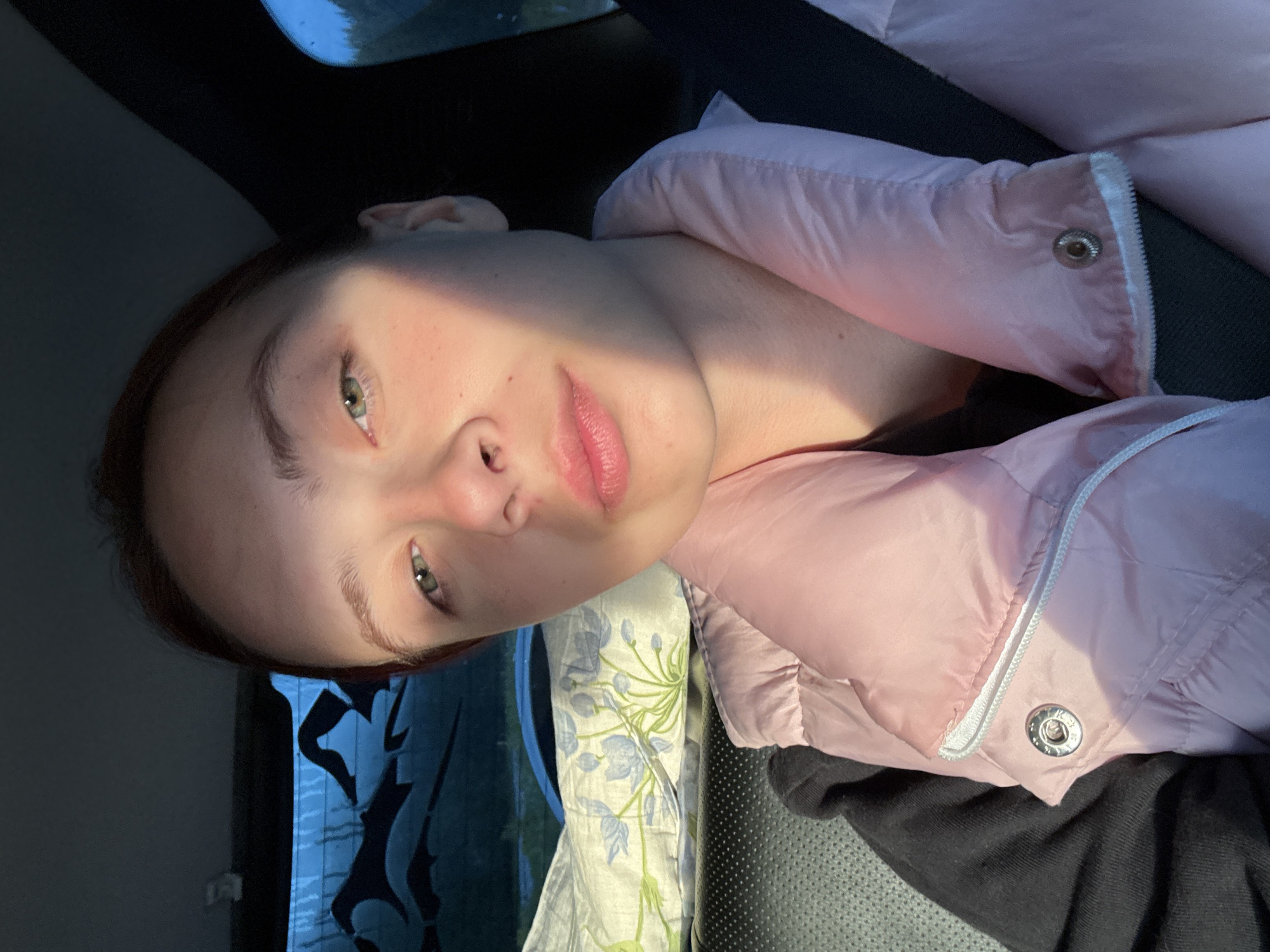 A young woman is lying down in what appears to be a car. She is wearing a pink puffer jacket and is resting on a pillow with a floral pattern. Sunlight is shining on her face. The background suggests the interior of a vehicle and a glimpse of an outdoor environment, possibly a roadside scene. There is no direct indication of the location being Прикамский, Russia, based solely on the visual elements of the image.