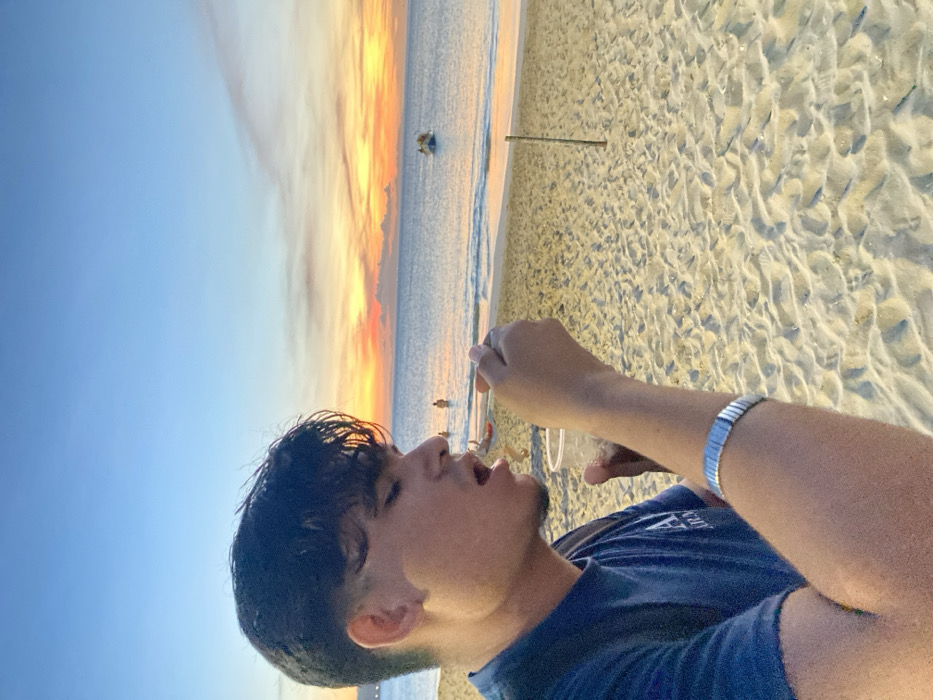 A young man drinks from a clear plastic cup at the beach, his face in profile. The setting sun casts a warm glow on the sky and the sand. The beach is textured with ripples and footprints. The ocean extends to the horizon, with hints of surfers in the distance. He wears a dark-colored shirt and a silver watch. His hair is short and dark.