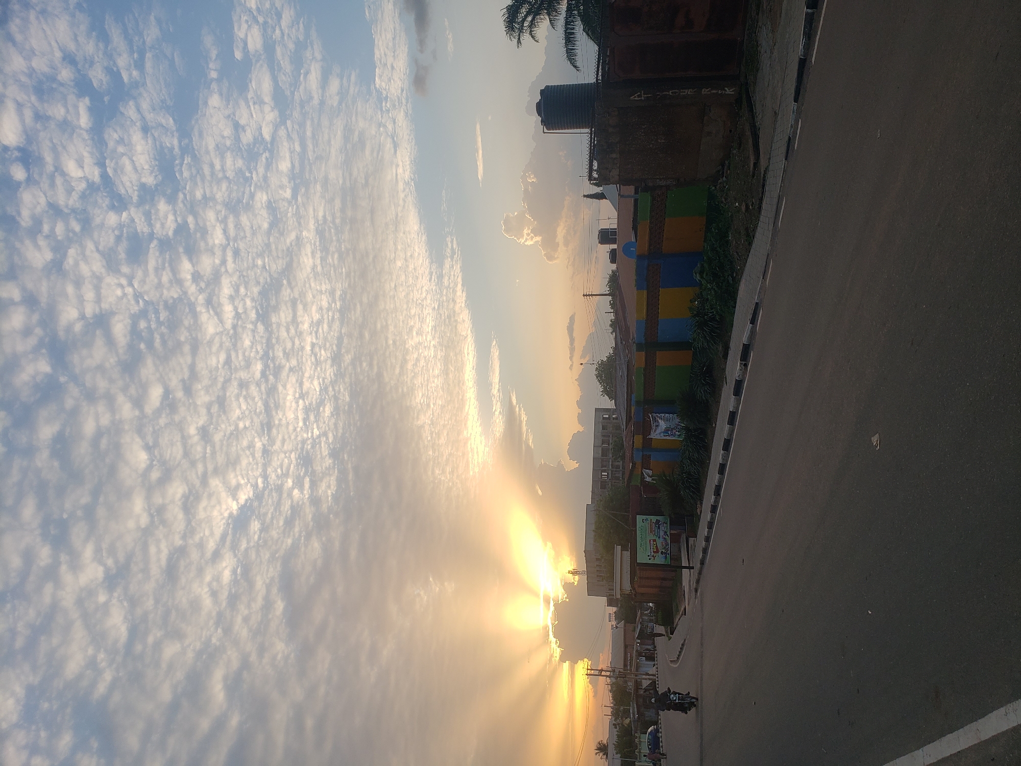 The image depicts a street scene in Narayi, Nigeria, at sunrise or sunset. The sky is filled with scattered clouds, with bright sunrays emanating from behind a bank of clouds on the left, casting a warm glow over the scene. A road runs diagonally through the frame, bordered by a sidewalk. On the right side of the road, there are several buildings, including a water tank and a wall decorated with colorful rectangular panels. A billboard with text and an image is also visible. On the left side of the road, there are more buildings and a few distant figures, including a person on a motorcycle. The overall impression is one of an urban environment bathed in the soft light of dawn or dusk.