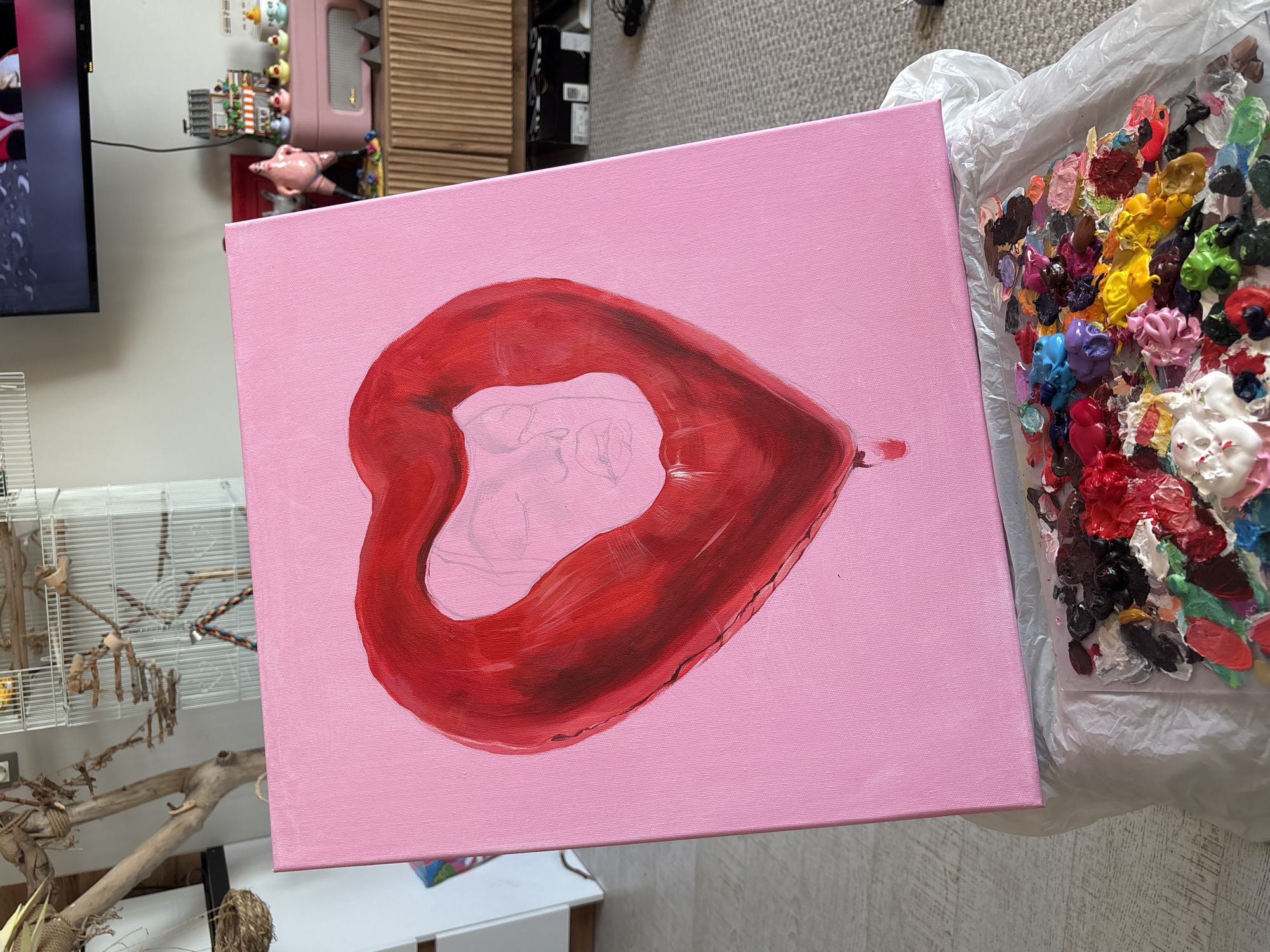 An overhead shot captures an ongoing painting project and its surroundings within what appears to be a creative personal space in Krasnoyarsk, Russia. The central subject is a rectangular canvas painted in a vibrant pink hue. On this canvas, a large, heart-shaped pair of red lips is in progress, with shades of crimson and darker reds giving it a shiny, almost balloon-like appearance. Within the unpainted white space inside the lips, a faint pencil outline of a person's head and upper body is visible, suggesting the next layer of the artwork.
To the right of the canvas, a disposable plastic palette is heavily laden with a multitude of paint blobs in various colors, including bright reds, blues, yellows, greens, purples, and whites, indicating an active painting session. The colors are both fresh and partially dried.
The background reveals parts of a room. On the left, a TV screen displays a portion of what looks like a person's profile. Below the TV and extending to the left, a white wire bird cage with wooden perches and a rope toy is visible, alongside a substantial, natural-looking wooden branch structure, likely part of a bird's habitat. Further into the background, above the bird cage, a pink retro-style radio or stereo is positioned on a surface, accompanied by several small, colorful decorative figurines, including what appears to be tiny ducks and a larger pink figure resembling a person in a festive outfit. The floor is covered with a light-colored, textured carpet. No visible text is clearly discernible on any of the objects.