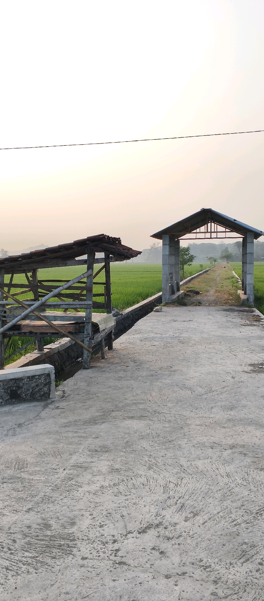 A concrete path leads through an open field of green rice paddies under a hazy sky, possibly at dawn or dusk. On the left, a rustic wooden structure with a tiled roof and bamboo supports sits beside the path, overlooking a narrow, dark channel of water. On the right, a more modern, gray stone archway stands as an entrance to the path, its roof also tiled and supported by a metal frame. The path appears to be a walkway or road, with some vegetation growing along its edges. The overall scene suggests a rural landscape in Japanan, Indonesia, likely a farming area.
