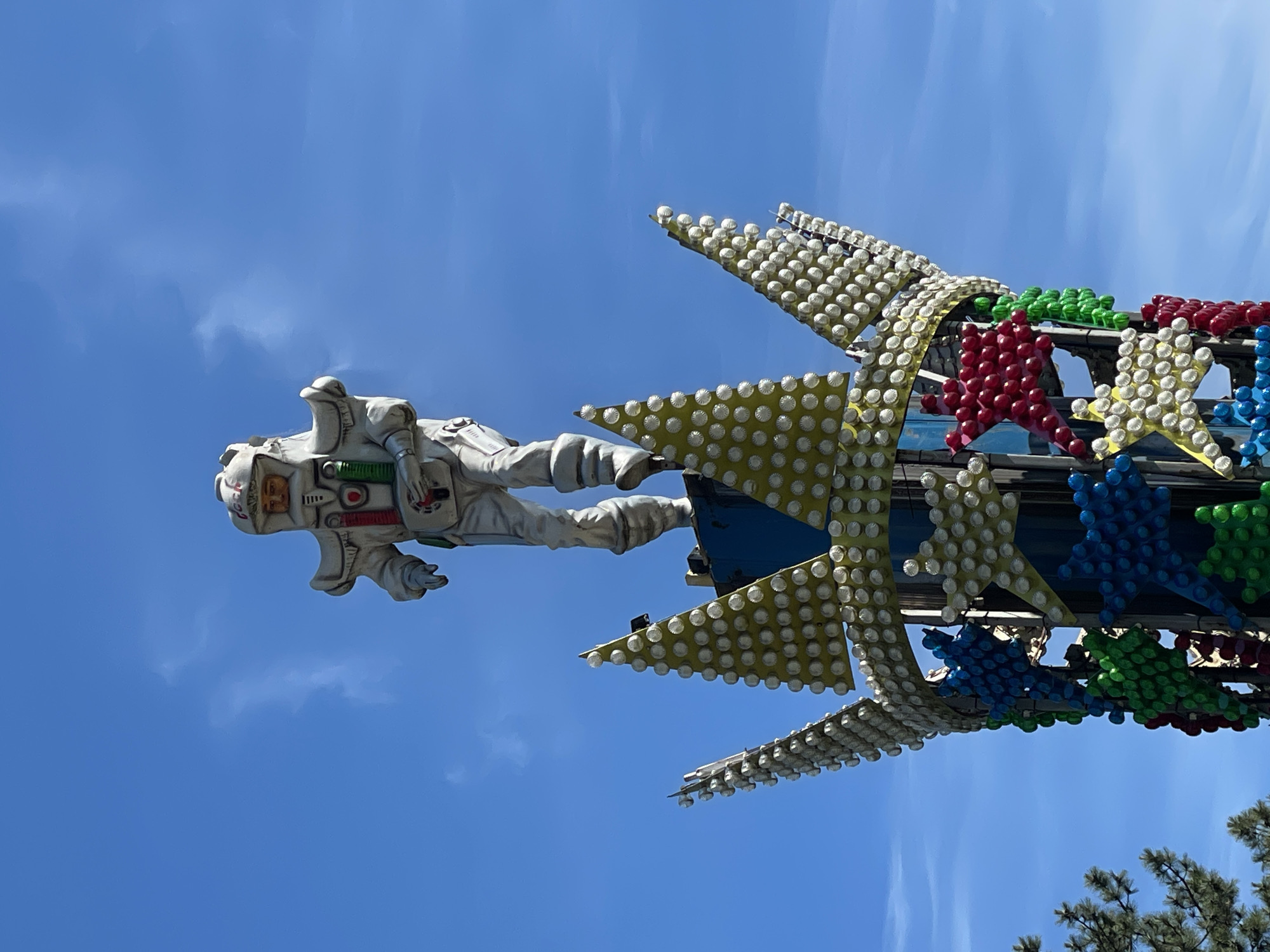 This image, captured in Sochi, Russia, features a large, detailed figure of an astronaut in a white spacesuit, appearing to float against a bright blue sky. The astronaut is positioned as if in zero gravity, with arms bent and legs slightly splayed. The suit includes various colored accents such as red and green details on the chest, a visible face behind the helmet's visor, and dark gloves.
To the right and below the astronaut, a colorful amusement park ride structure is prominently displayed. This structure is composed of numerous star-shaped elements, many adorned with arrays of white, round light bulbs set against a light yellow background. Other stars are solid red, green, or blue, also covered with similar light bulbs, suggesting a festive, light-up attraction. The ride appears to be in a static state.
The scene is set outdoors on a clear and sunny day, indicated by the vibrant blue sky with minimal, wispy white clouds. Pine tree branches are visible in the bottom right corner, suggesting the presence of natural foliage around the park. There is no discernible activity or event taking place; the image is a still shot of the attraction. While there are symbols and patches on the astronaut's suit, no clear or legible text is visible on any of the subjects. The overall context points to a space-themed attraction at an amusement park or fairground.
