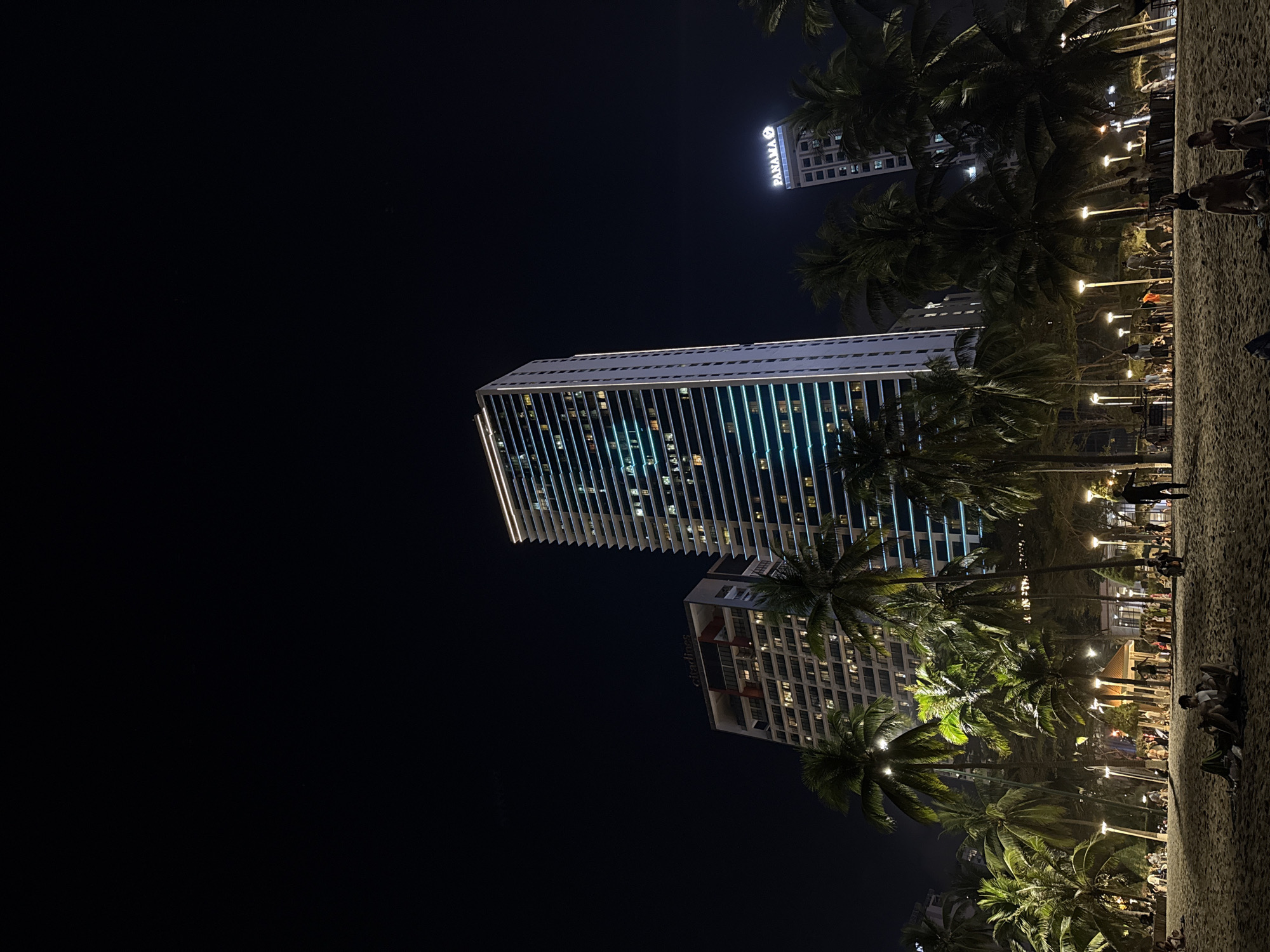 The image depicts a nighttime scene on a beach in Nha Trang Ward, Vietnam. A tall, modern skyscraper with illuminated windows dominates the view, positioned next to an illuminated Citadines building. A secondary building in the distance has "PANAMA" displayed on the front. The beach is populated with palm trees and illuminated by streetlights and lampposts. People are present on the beach. The overall environment is dark, with the primary sources of light originating from the buildings, streetlights, and surrounding areas.