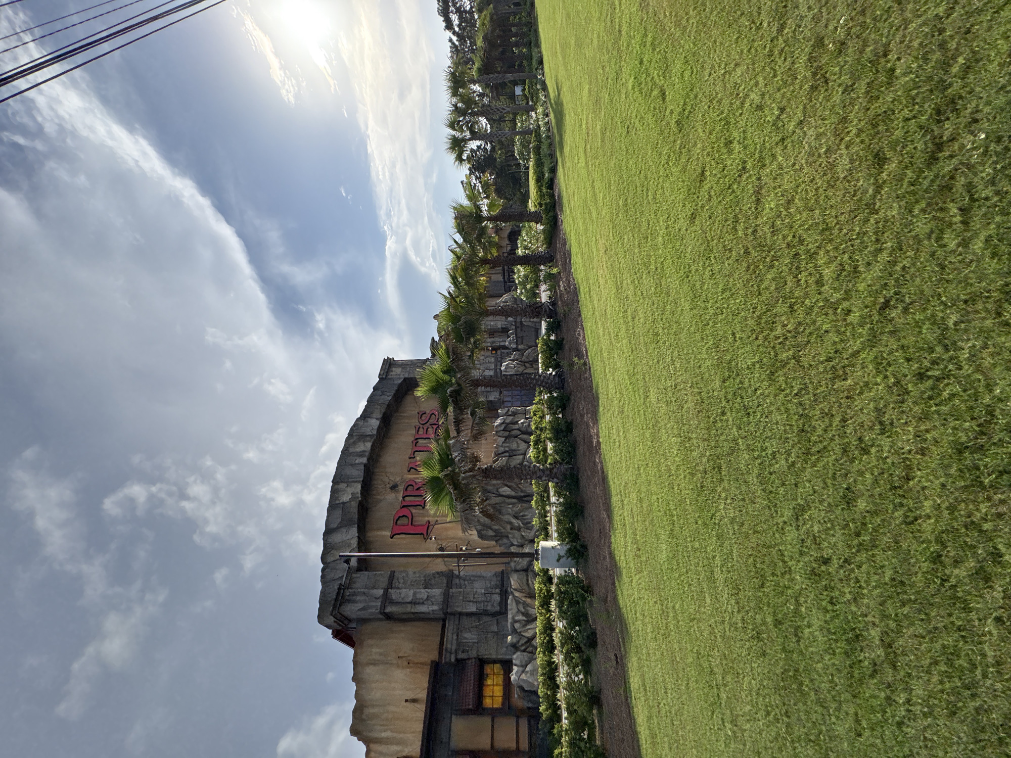 The image shows the Pirates Island Adventure Golf course in Panama City Beach, Florida. The course is located next to a green field, and several palm trees are positioned along the edge. The structure of the building with the name "PIRATES" in red lettering can be seen as well. The sky is cloudy with some sunshine breaking through. In the upper left, power lines can be seen.