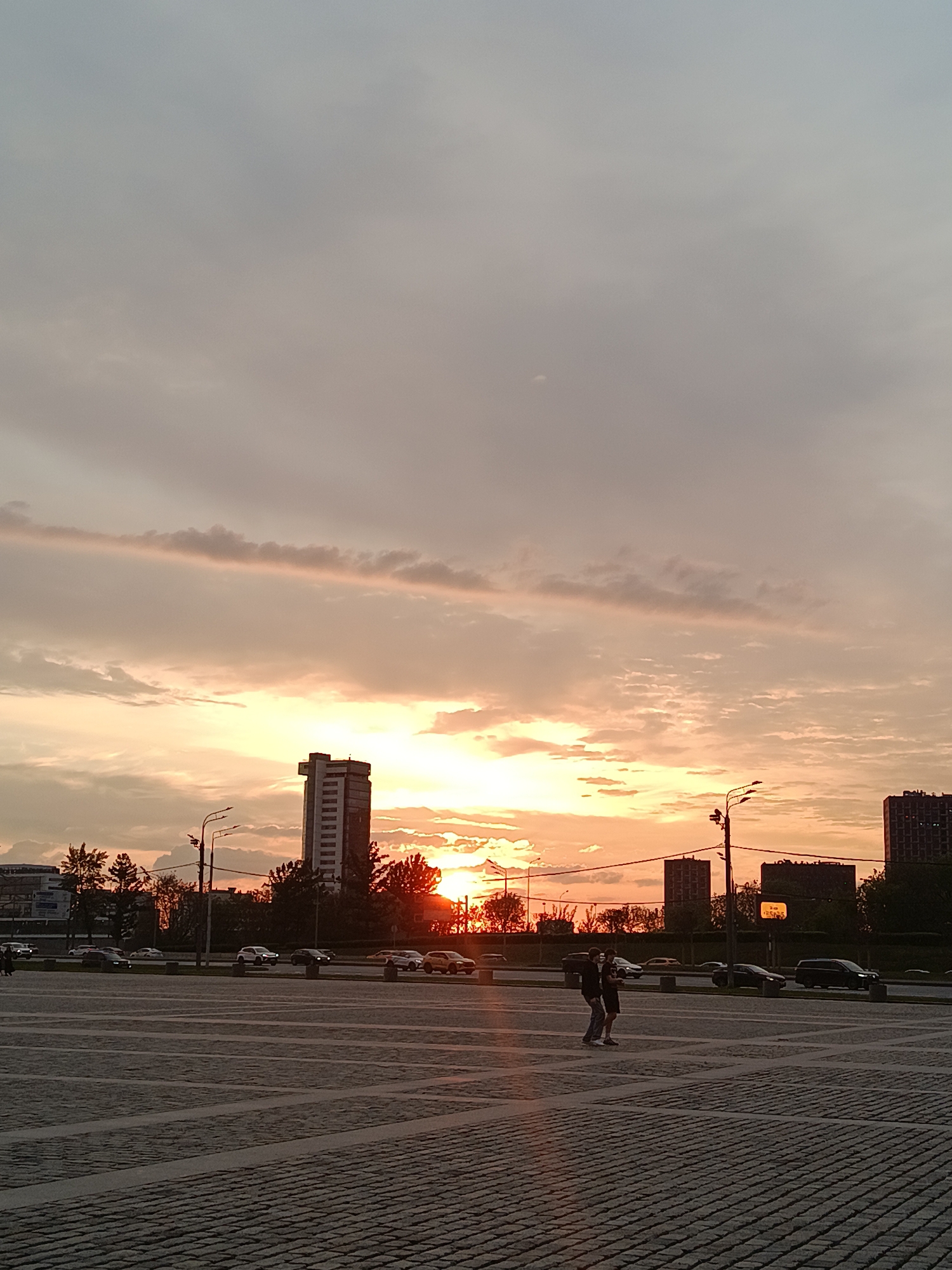 A low-angle long shot reveals a sprawling urban plaza bathed in the warm hues of a sunset. The sky, dominated by layered clouds tinged with pink and orange, sets the backdrop for a cityscape silhouetted against the bright horizon.
The plaza itself is paved with grey stones, arranged in a grid-like pattern. Two individuals are captured mid-stride, walking across the plaza, their figures defined by the backlighting.
Beyond the plaza, a multi-lane road teeming with vehicular traffic separates the foreground from a row of buildings of various heights. A prominent skyscraper, situated on the left, is dwarfed by the expansive sky. Streetlights stand as sentinels along the road.
This is Moscow, Russia.