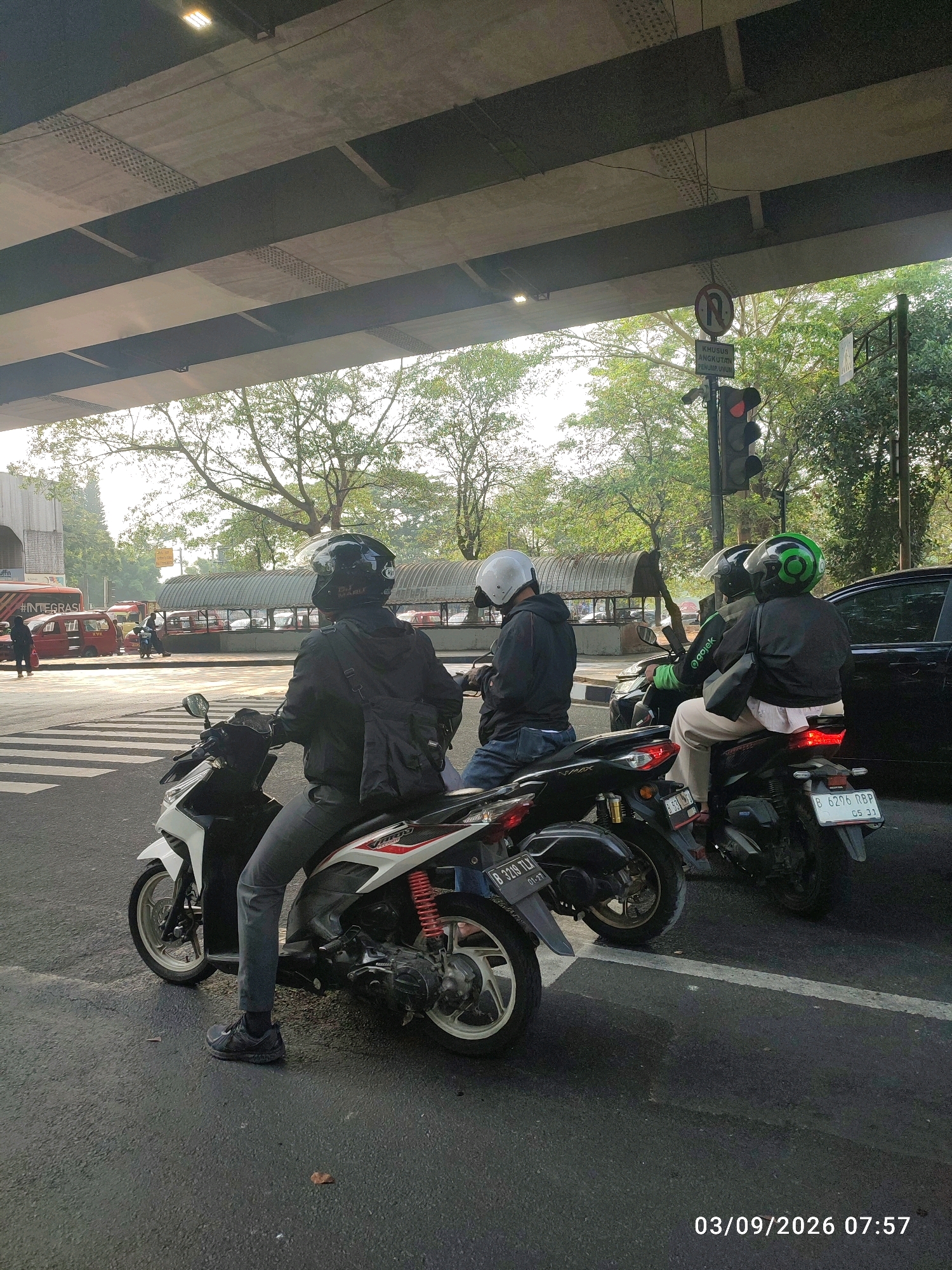 The image captures a street scene in East Jakarta, Indonesia, beneath an elevated roadway. Several motorcycles are stopped at what appears to be a traffic light. The rider in the foreground, facing away from the camera, wears a black helmet, black jacket, and gray pants, with a black bag slung over their shoulder. To the right, two individuals are on another motorcycle; the rear rider wears a green helmet and a jacket with "Gojek" branding, a ride-hailing service prominent in Indonesia. The front rider wears a black helmet. The environment includes trees, traffic signals, and other vehicles in the background, suggesting an urban setting. The date and time stamp at the bottom right corner reads "03/09/2026 07:57".