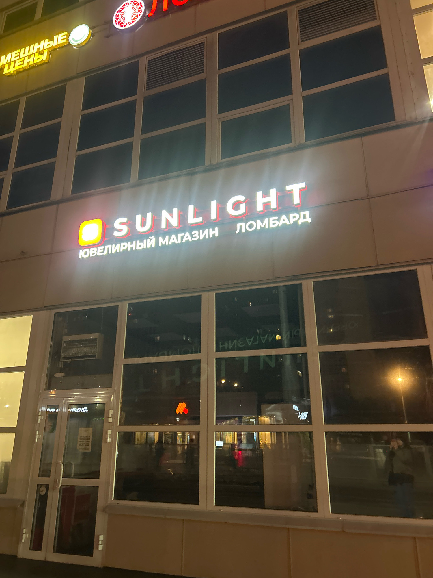 The image shows the exterior of a Sunlight jewelry store and pawnshop at night in Moscow, Russia. The store has large glass windows and a brightly lit sign that reads "SUNLIGHT" in large white letters, with "Ювелирный магазин" (Jewelry store) and "ЛОМБАРД" (Pawnshop) written below in smaller letters. There are also other signs on the building, including one that says "МЕЧНЫЕ ЦЕНЫ" (Bargain prices). Inside the store, reflections in the windows show people and other businesses. A street lamp is visible in the background. The overall impression is that of a modern commercial establishment operating in an urban environment.