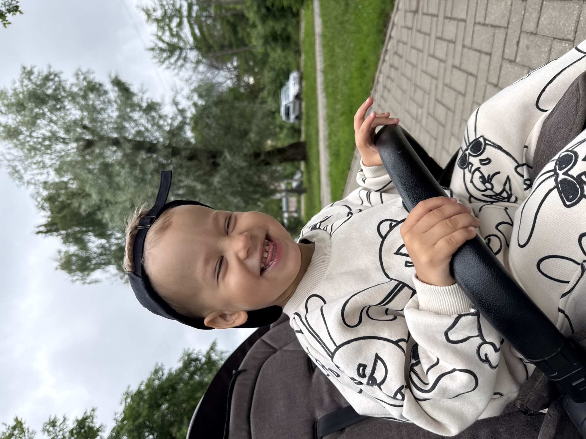 A young boy wearing a white sweatshirt with a black cartoon print and a black baseball cap sits in a stroller, holding onto the stroller's handle. He has his eyes closed and is smiling broadly, showing his teeth. The stroller is on a paved walkway in what appears to be a park or residential area, with green grass and trees in the background. A silver car is visible in the distance. The sky is overcast.