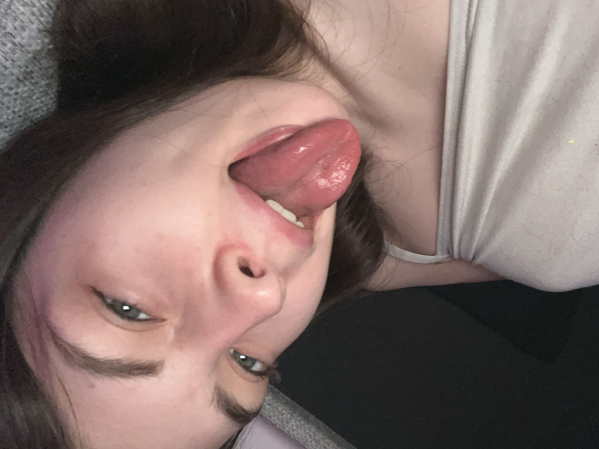 A woman lies supine with her head at an inverted angle, protruding her tongue outward. She wears a light-colored tank top. Her eyes are open, and her hair is dark. The environment appears to be a neutral-toned interior setting, possibly a residential space, resting on a dark surface. The location is unknown.