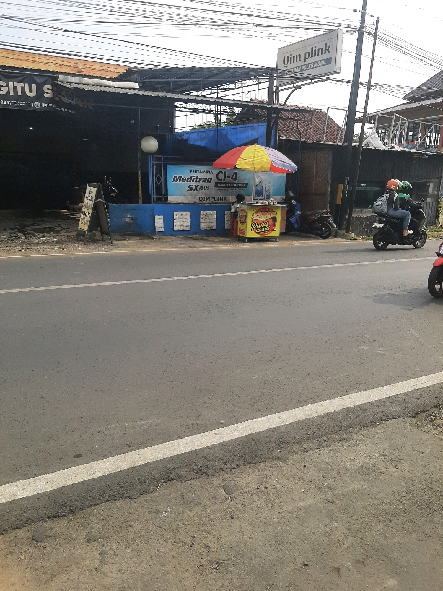 A roadside view captures a car wash and detailing service named "Qim plink" in Purwanegara, Indonesia. A sign above the building advertises "Cuci dan Poles Mobil" (Car Wash and Polish). In the foreground, a street runs alongside the establishment. A yellow and red umbrella shades a food cart with the branding "Pukis Abadi" (Eternal Pukis). Two individuals on a motorcycle, wearing helmets and backpacks, are seen passing by. Another motorcycle is parked near the food cart. The car wash building has a dark blue facade, and a white banner with text and a logo is displayed. Electrical wires crisscross the sky above.