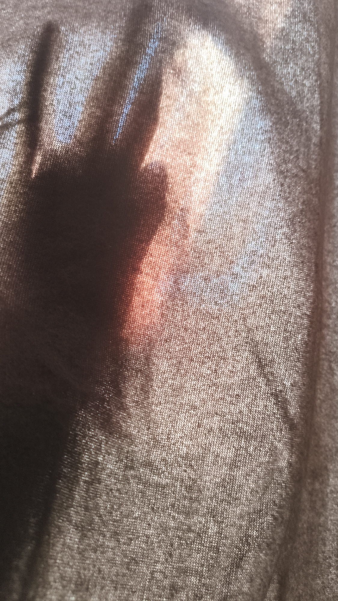 A hand shadow is projected onto a textured fabric, likely a curtain or other sheer material. The silhouette of the hand is dark and covers a significant portion of the image's left side. Light filters through the fabric, creating an orange glow. The background appears to be light, possibly daylight. The details necessary to determine the location as Nowy Tomyśl, Poland, are not present in the image.