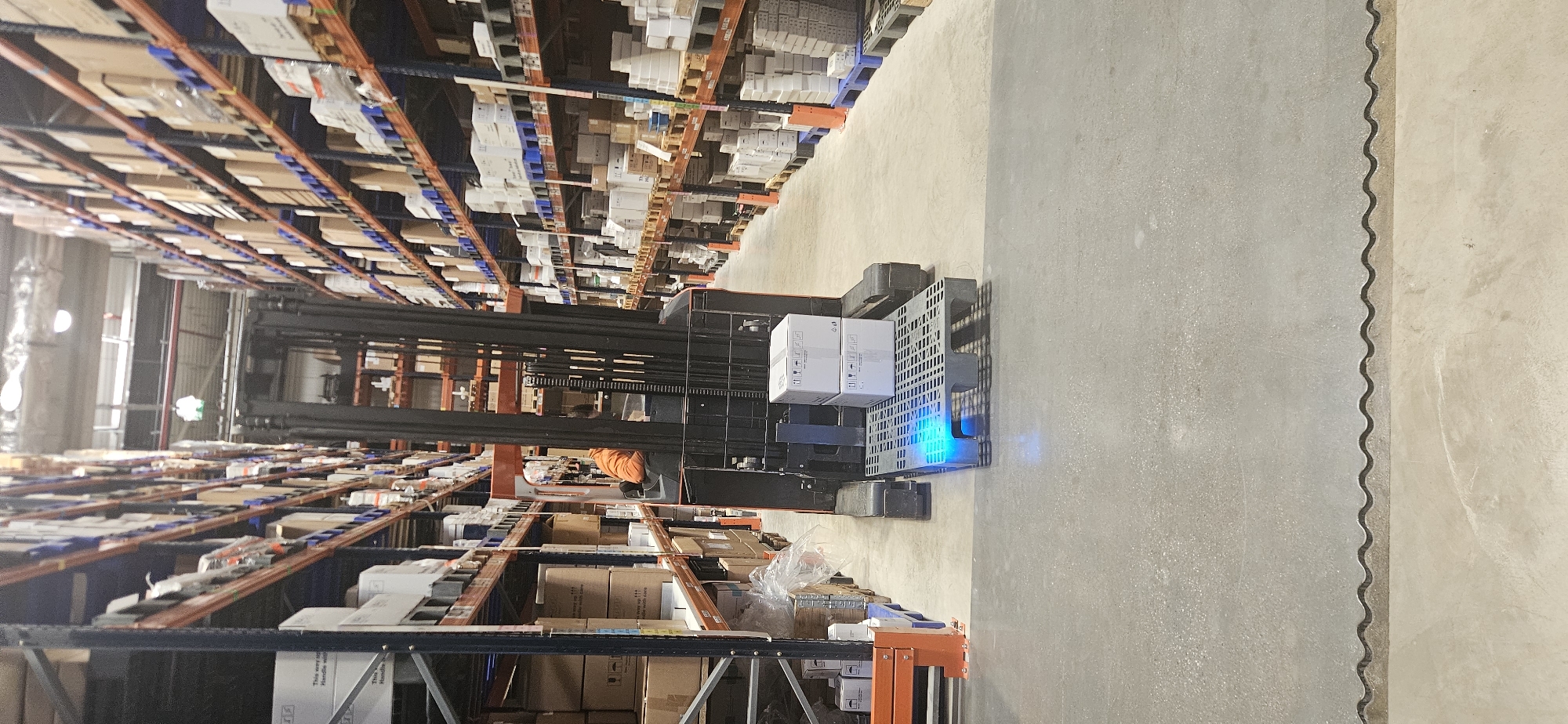 A tall forklift, carrying two white boxes, stands in a narrow aisle between tall warehouse storage racks. The racks are densely filled with boxes of various sizes and colors. The forklift has a blue light emitting from its base. An individual in orange clothing appears to be operating the forklift. The floor is concrete. The location is potentially a warehouse in Plovdiv, Bulgaria, given the request.