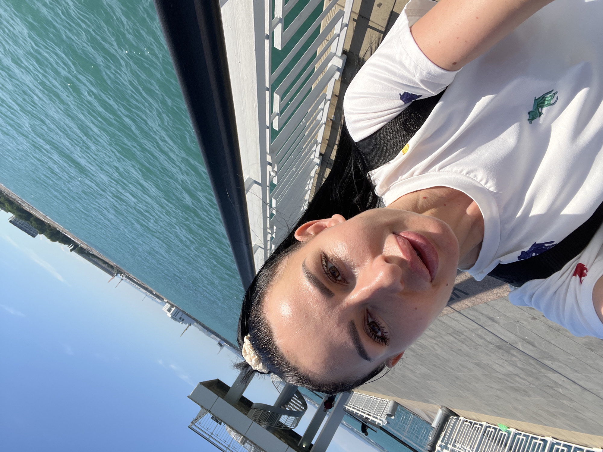 The image is a selfie of a woman with long black hair, wearing a white t-shirt with colorful polo horse logos, and a black backpack. She is on a pier or boardwalk, with the sea to her right and a walkway to her left. In the background, there is a blue sky, the turquoise sea with gentle waves, and some buildings and structures visible along the coastline, suggesting an urban seaside location. The woman is looking at the camera with her mouth slightly open. The lighting indicates it is daytime, with sunlight casting shadows. Based on the visual cues of the architecture and the sea, this location is likely the Burgas Sea Garden in Burgas, Bulgaria, specifically near the pedestrian bridge and the beach.