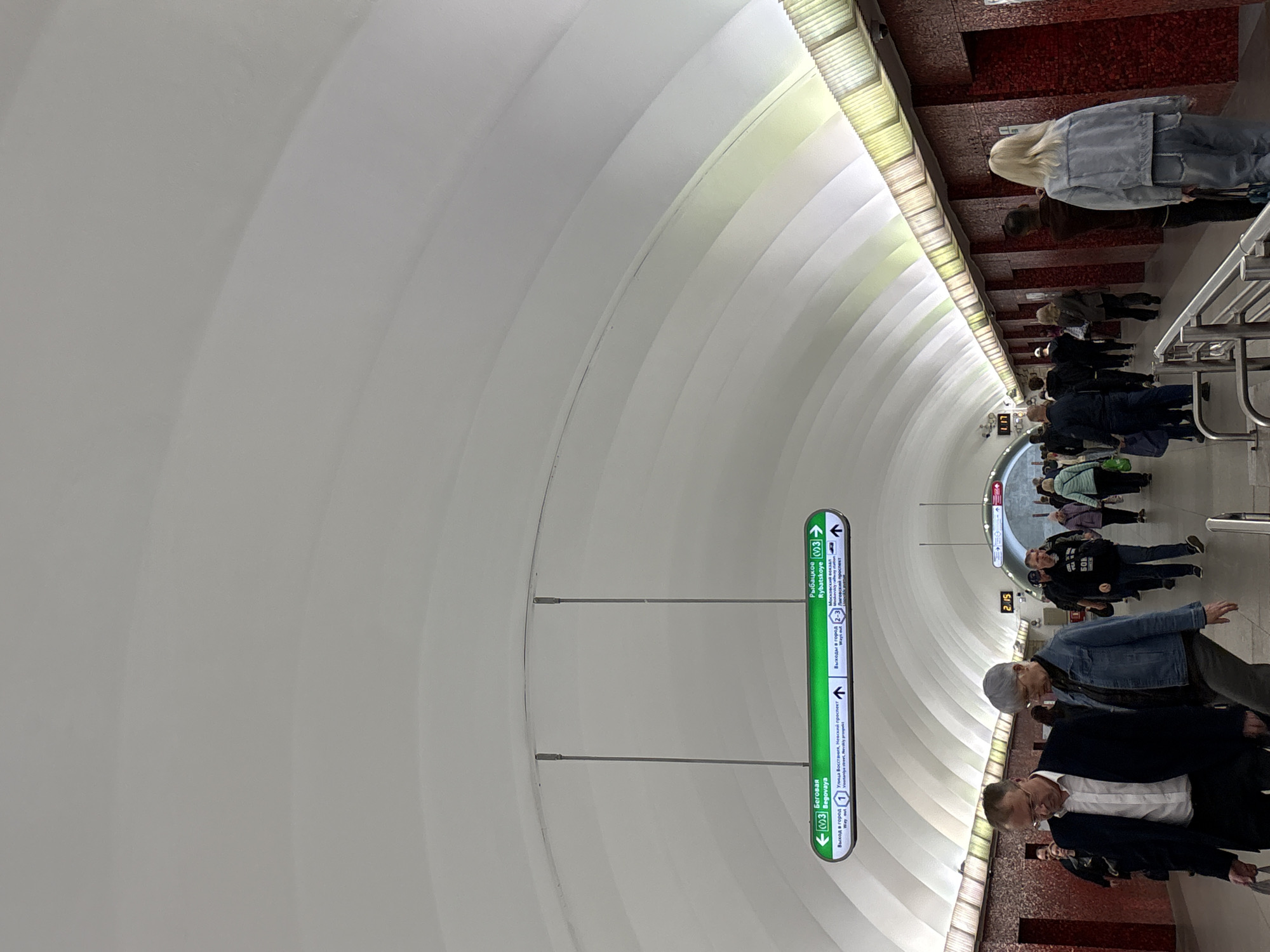 The scene is inside a metro station in Saint Petersburg, Russia. The station features a curved, white ceiling. People are walking toward the station exit. There is a sign indicating directions to the "Begovaya" (exit 1) and "Rybatstkoye" (exits 2-3) destinations and Moscow station. The sign also mentions "Ulitsa Vosstaniya" and "Nevsky Prospekt."