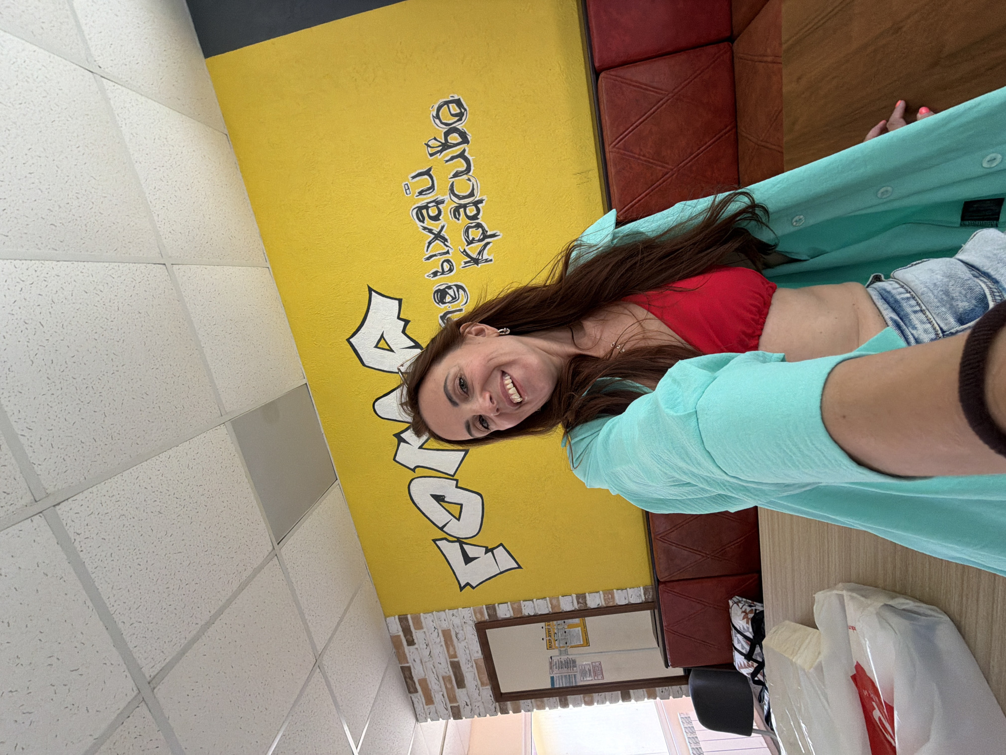A woman with long brown hair, wearing a red bikini top and a teal open button-up shirt, takes a selfie indoors. She is smiling at the camera. Behind her, a bright yellow wall features graffiti-style text in black and white. The text appears to be in Cyrillic script. Red banquettes are visible along the wall. A light-colored ceiling with acoustic tiles is overhead. To the left, a brick-patterned wall is visible, with a framed mirror and some papers hanging on it. A wooden table is in the foreground. The overall setting suggests a casual indoor establishment. This location is in Birobidzhan, Russia.