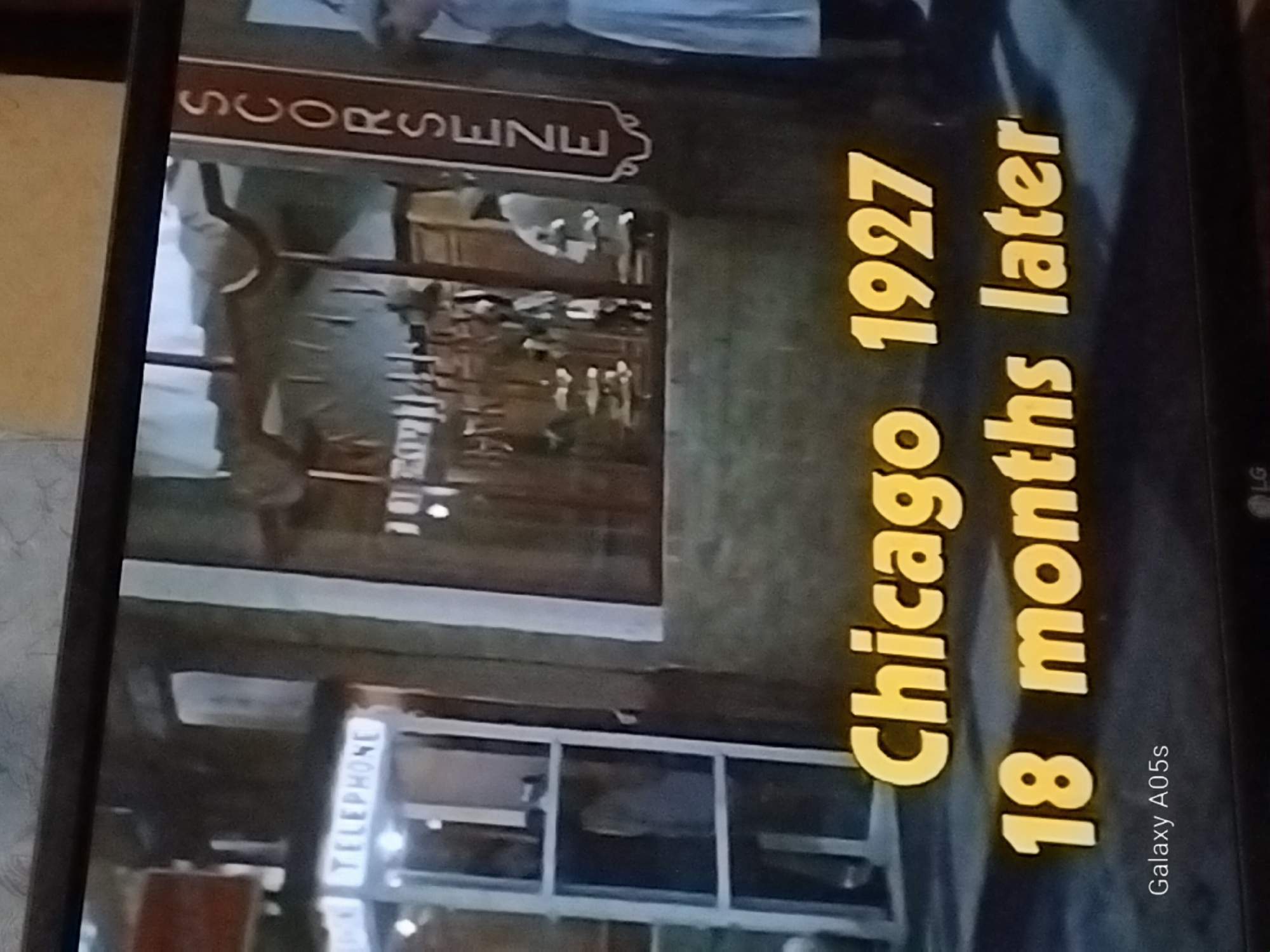 The image shows a scene from a movie or television show, possibly a period piece, with text overlays. In the background, a vintage telephone booth is visible with the word "TELEPHONE" illuminated above it. To the right, a vertical sign with stylized lettering reads "SCORSENZE". The foreground features large yellow text stating "Chicago 1927" and below it, "18 months later". There are no discernible elements in the image that indicate the location is Druzhba, Russia; the text "Chicago 1927" strongly suggests an American setting. The scene appears to be set in an urban environment from the early 20th century.