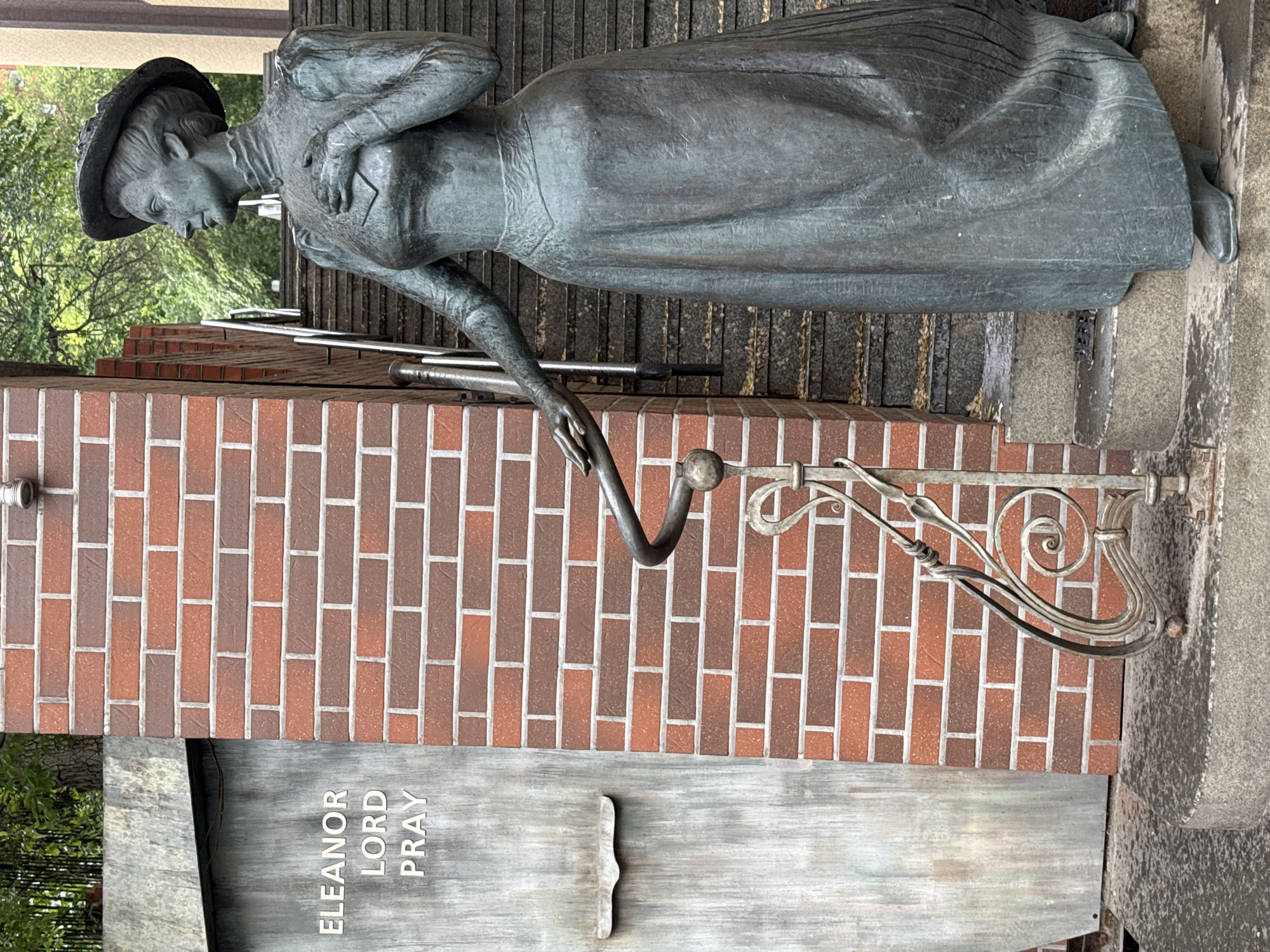 The image displays a vertical outdoor shot of a statue commemorating Eleanor Lord Pray. The statue, made of dark, patinated bronze or similar metal, depicts a woman in a long dress and a hat, characteristic of the late 19th or early 20th century. She is shown in a walking or contemplative pose, leaning slightly forward with her right hand gently resting on an ornate, dark metal handrail that features decorative scrolls. Her left hand is placed across her chest. The woman's head is tilted downwards, suggesting a pensive or observant expression.
The statue is situated alongside a brick wall constructed from reddish-brown and darker bricks. Behind the statue and the wall, a set of stone stairs ascends, with lush green foliage from trees or bushes visible in the background, indicating an outdoor, possibly park-like or urban setting with natural elements.
At the base of the brick wall, beneath the statue, a dark, weathered rectangular plaque is affixed. Vertically inscribed on this plaque in capital letters is the name "ELEANOR LORD PRAY." To the right of the name, a decorative, vertical, metal bracket-like embellishment is attached to the plaque.
The lighting appears soft and diffused, suggesting daytime under overcast conditions or in shade, as there are no harsh shadows. The overall scene evokes a sense of historical commemoration and quiet dignity in Vladivostok, Russia.