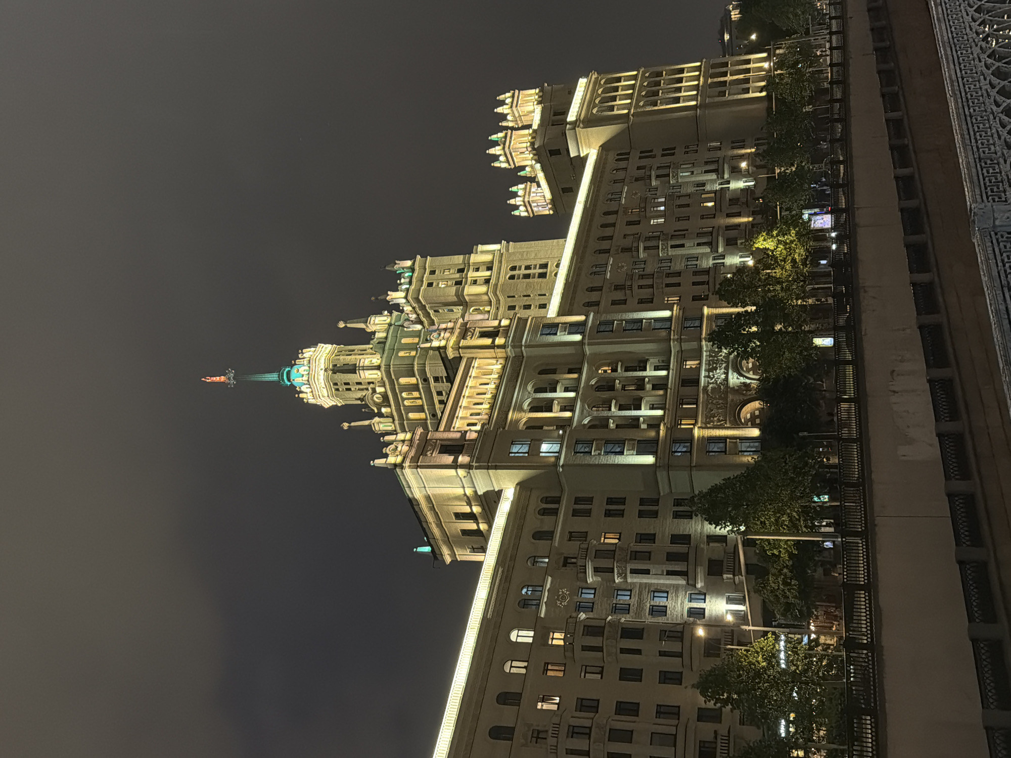 At night, a brightly lit Stalinist skyscraper rises against a dark sky. The architecture features multiple tiers, ornate detailing, and a spire topped with a star. A section of a nearby building is visible on the right side. Trees and streetlights are along the base of the building. The location is Перовское сельское поселение, Ukraine.