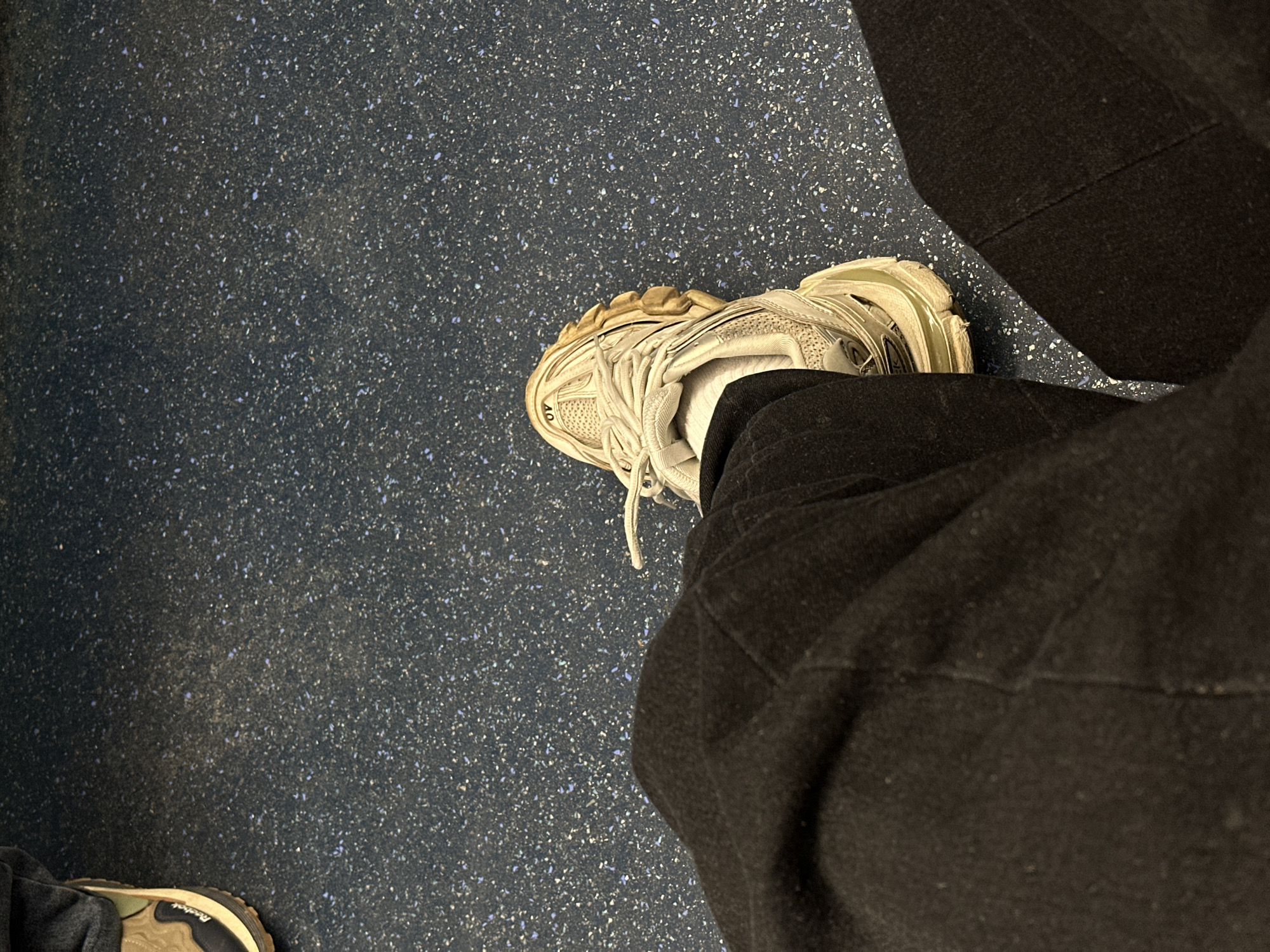 The image provides a downward, high-angle view of two individuals' legs and feet on a speckled dark blue floor. The main subject in the foreground and center-right is a leg clad in dark, possibly black, trousers or sweatpants, with a light-colored, chunky athletic shoe (likely white or off-white) visible. The shoe appears well-worn, with dirt visible on its sole and parts of the upper. The number "40" is distinctly visible on the toe area of this shoe, indicating a European shoe size.
In the bottom-left corner, a portion of a second leg and foot is visible. This individual is also wearing dark trousers and a different athletic shoe, featuring tan/green and black elements on its sole. The brand name "Reebok" is partially discernible on the side of the sole of this shoe.
The floor is a dark blue with numerous small, irregularly shaped speckles of various lighter colors, including white, light blue, and grey. This type of flooring is commonly found in public spaces, such as public transport vehicles or stations, or certain institutional settings. The overall scene suggests an indoor environment. Given the context, this scene is located in Moscow, Russia. The time of day and weather are not discernible. No particular activity beyond being present in the space is evident. The image does not convey any specific emotions.