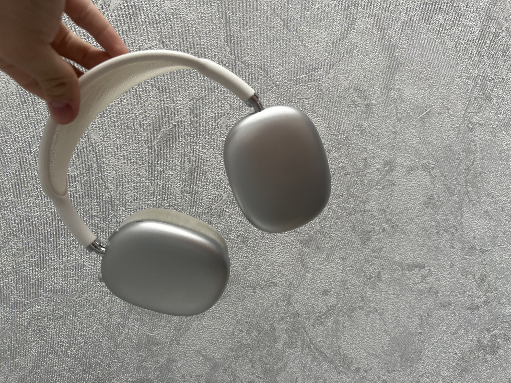 A person's hand holds the headband of a pair of silver and white over-ear headphones against a textured gray background. The headphones are positioned at an angle, with the earcups dangling downwards. The background appears to be a wall with a raised, ribbed texture. There are no other objects or individuals visible in the frame.