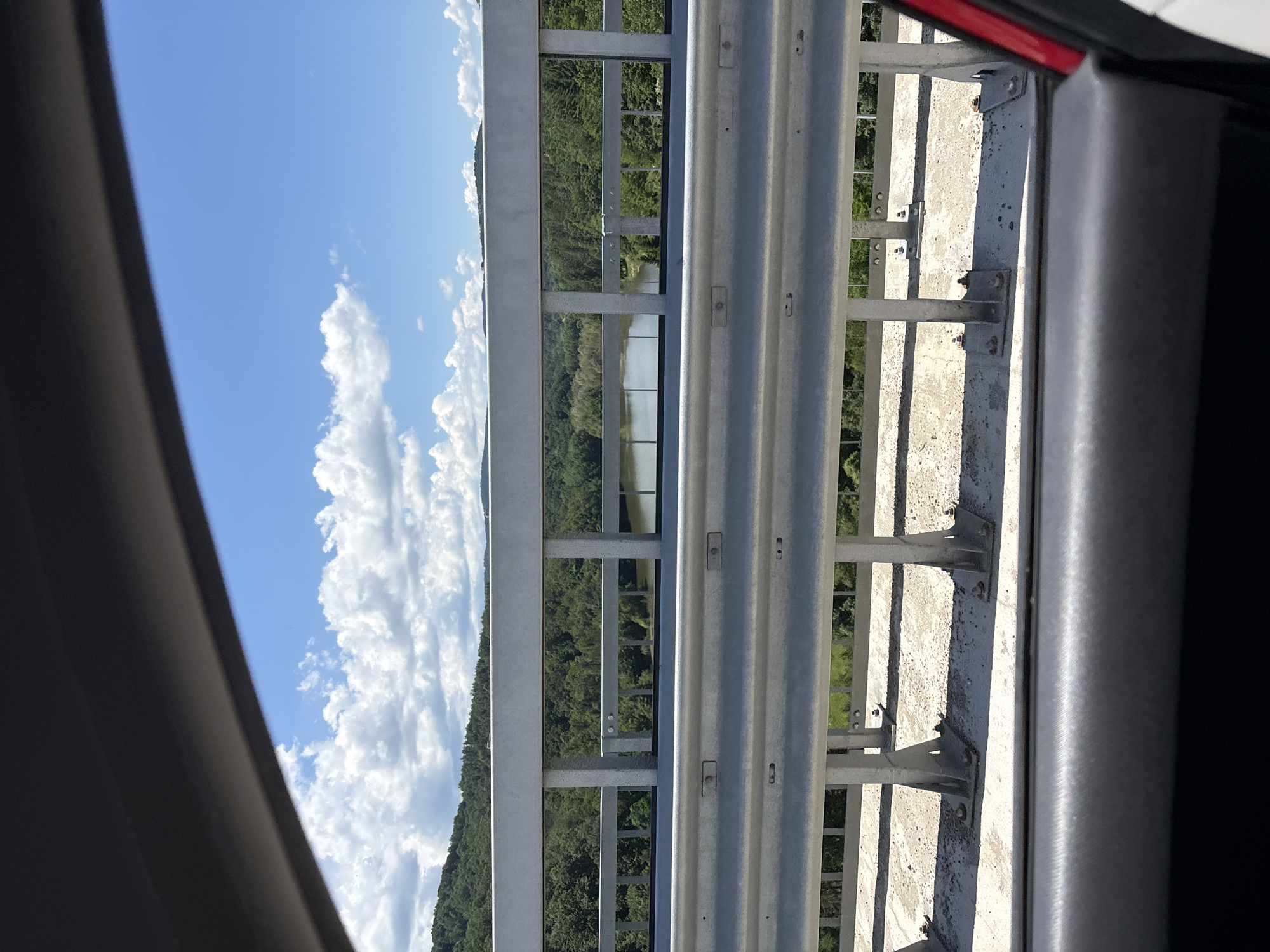A view from a car window shows a wide shot of a landscape on a clear, sunny day. In the foreground, a grey metal guardrail is visible, along with part of the car's interior. Beyond the guardrail, a body of water is seen, surrounded by lush green trees and rolling hills. The sky is a vibrant blue with scattered white clouds. The scene appears to be from a bridge overlooking a natural landscape, potentially a lake or river, in or near Gabrovo, Bulgaria.