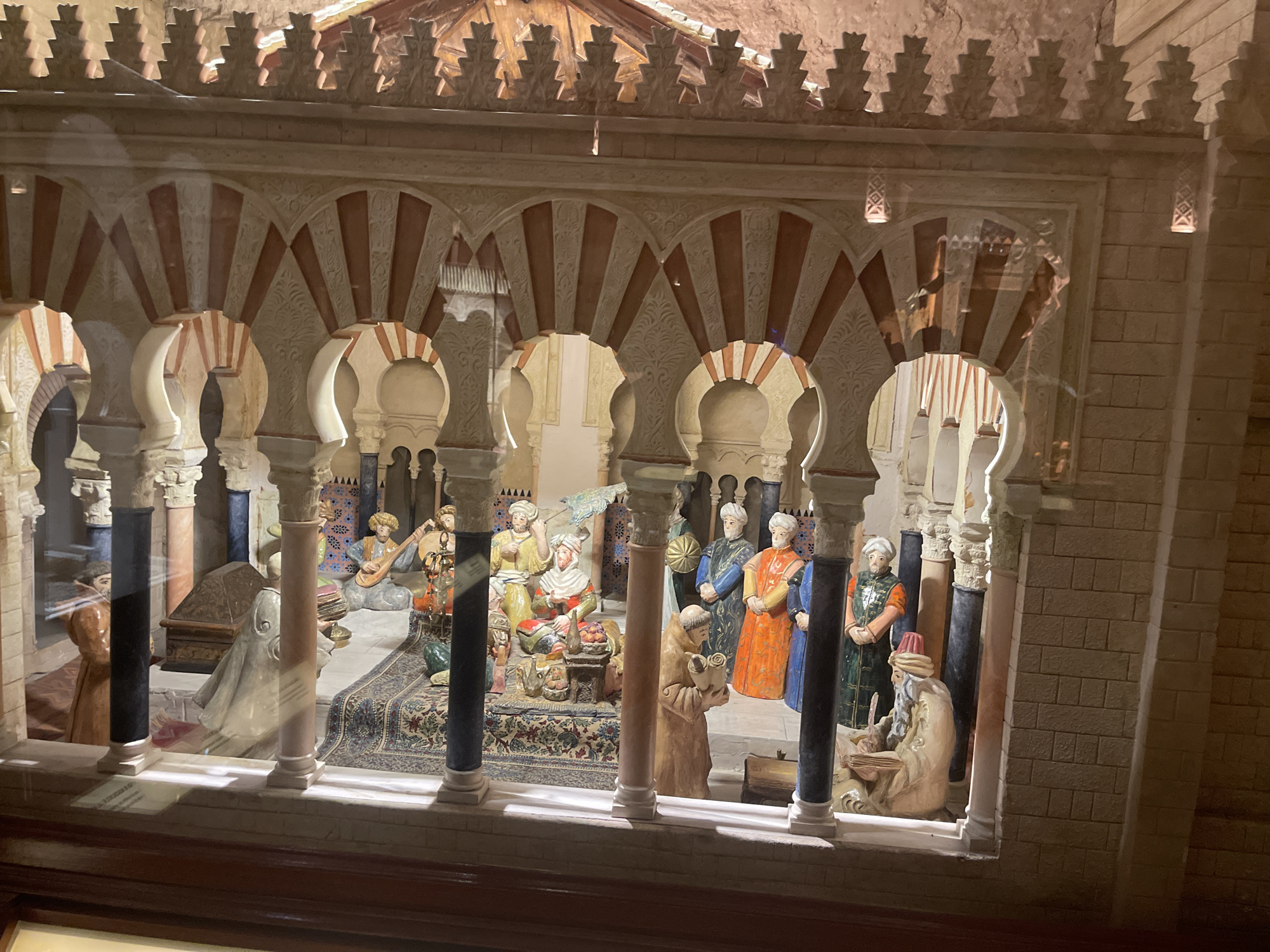 This is a diorama of the Mezquita-Cathedral of Córdoba, Spain, located inside the monument. The diorama depicts a scene from Islamic Cordoba, likely representing the caliphate era. It features a courtyard with characteristic horseshoe arches and columns. Several figures, dressed in historical attire, are arranged within the courtyard. Some figures are seated on the floor, one is playing a lute, and others are standing. A patterned rug is laid out on the floor. The diorama is encased in glass and illuminated, with a visible reflection on the glass surface. The background architecture of the diorama replicates the stonework and decorative elements found in the Mezquita.