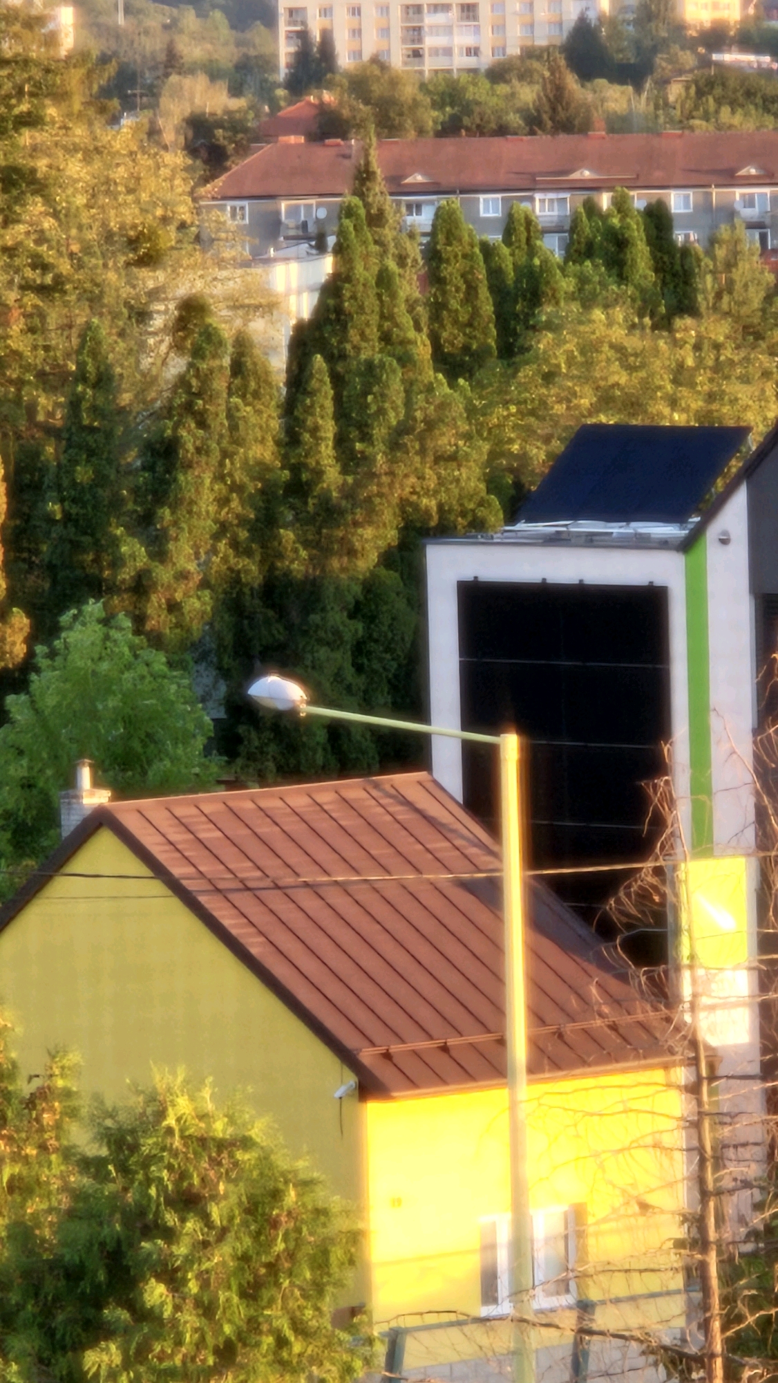 The image captures a view of a residential area, likely in Prešov, Slovakia, bathed in the warm glow of late afternoon or early morning sunlight. In the foreground, a bright yellow house with a reddish-brown tiled roof is visible, partially obscured by lush green trees. Adjacent to the house is a modern building featuring black solar panels on its facade and a distinctive green stripe. A white streetlamp extends across the frame, its light fixture a prominent white oval. In the middle ground, a row of older buildings with red-tiled roofs and white-framed windows can be seen, interspersed with dense evergreen trees. Further in the background, a larger, light-colored apartment building dominates the skyline, suggesting an urban or suburban setting. The overall impression is one of a peaceful neighborhood, with a blend of older and newer architectural styles set against a backdrop of natural foliage.