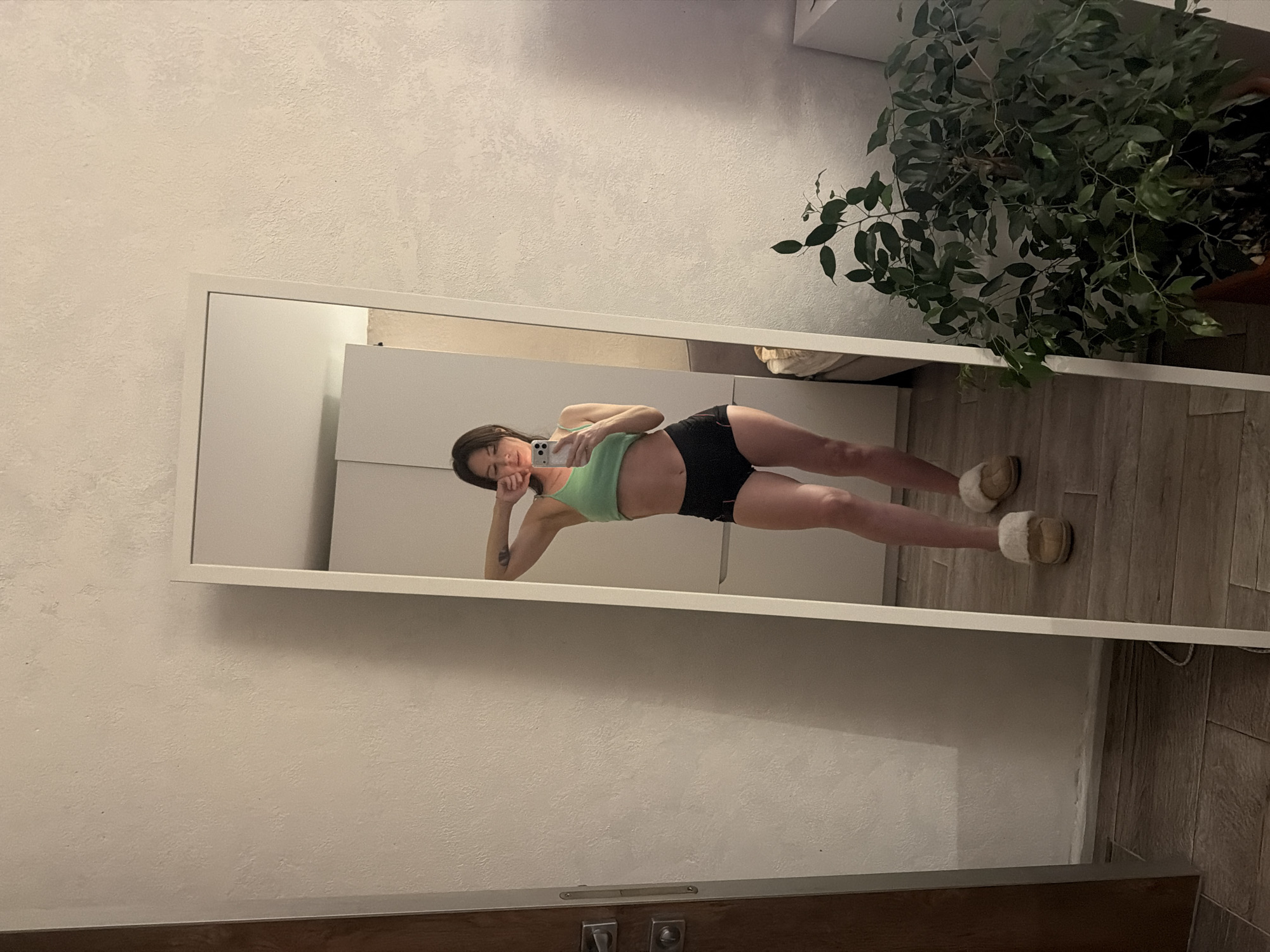 A mirror reflects a person posing in a green top and black shorts. The person is holding a phone, apparently taking a selfie. The surroundings include a plant, a white wall, and a wooden floor. It's unclear if the location is Perm, Russia.