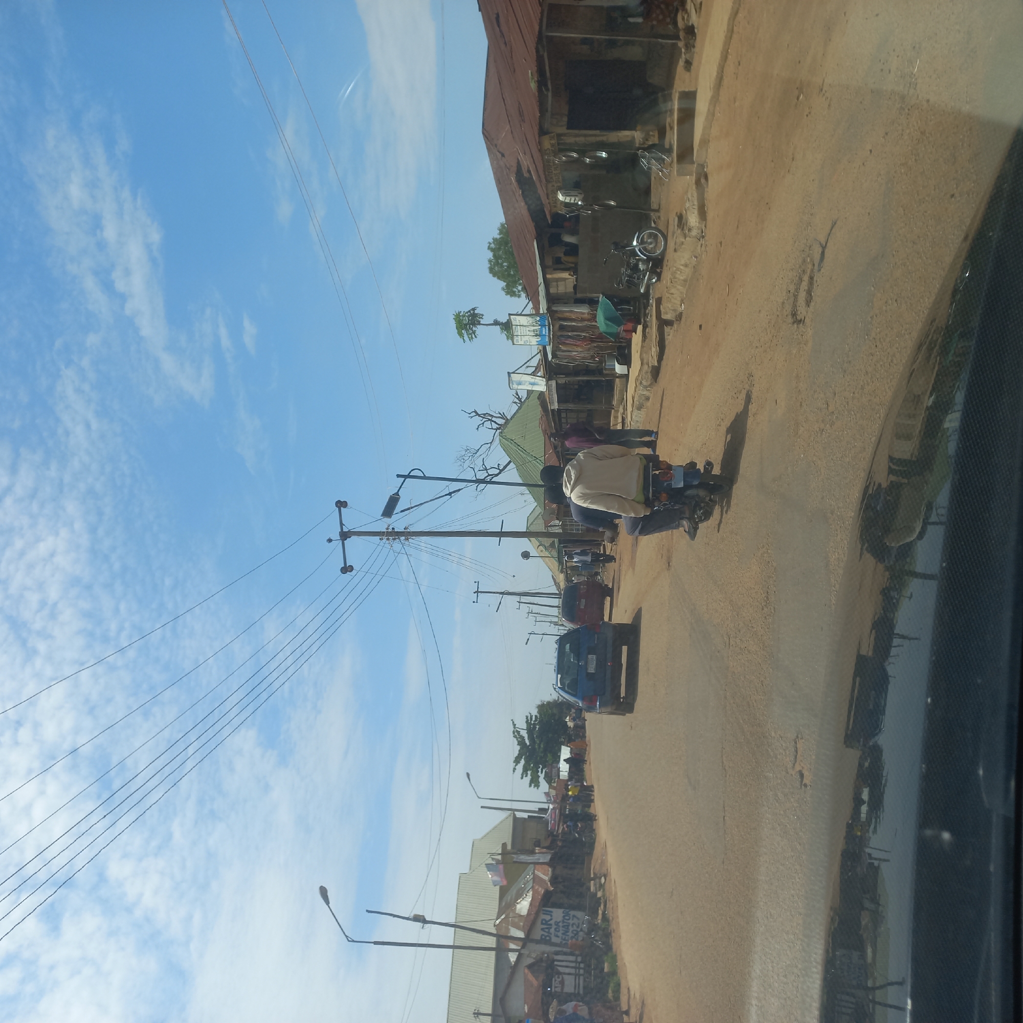 The image depicts a street scene in Mangu Town, Nigeria. The road is unpaved and dusty, with a few potholes visible. On either side of the road are various buildings, many with corrugated iron roofs, suggesting a commercial or residential area. Some of these buildings appear to be shops or market stalls. Several vehicles are present, including a blue SUV, a red car, and a motorcycle with two passengers. A few individuals are walking or standing around, observing the surroundings. The sky is blue with scattered clouds, and there are numerous electrical wires strung across poles, indicating the presence of power infrastructure. The overall impression is of a bustling, everyday scene in a Nigerian town.