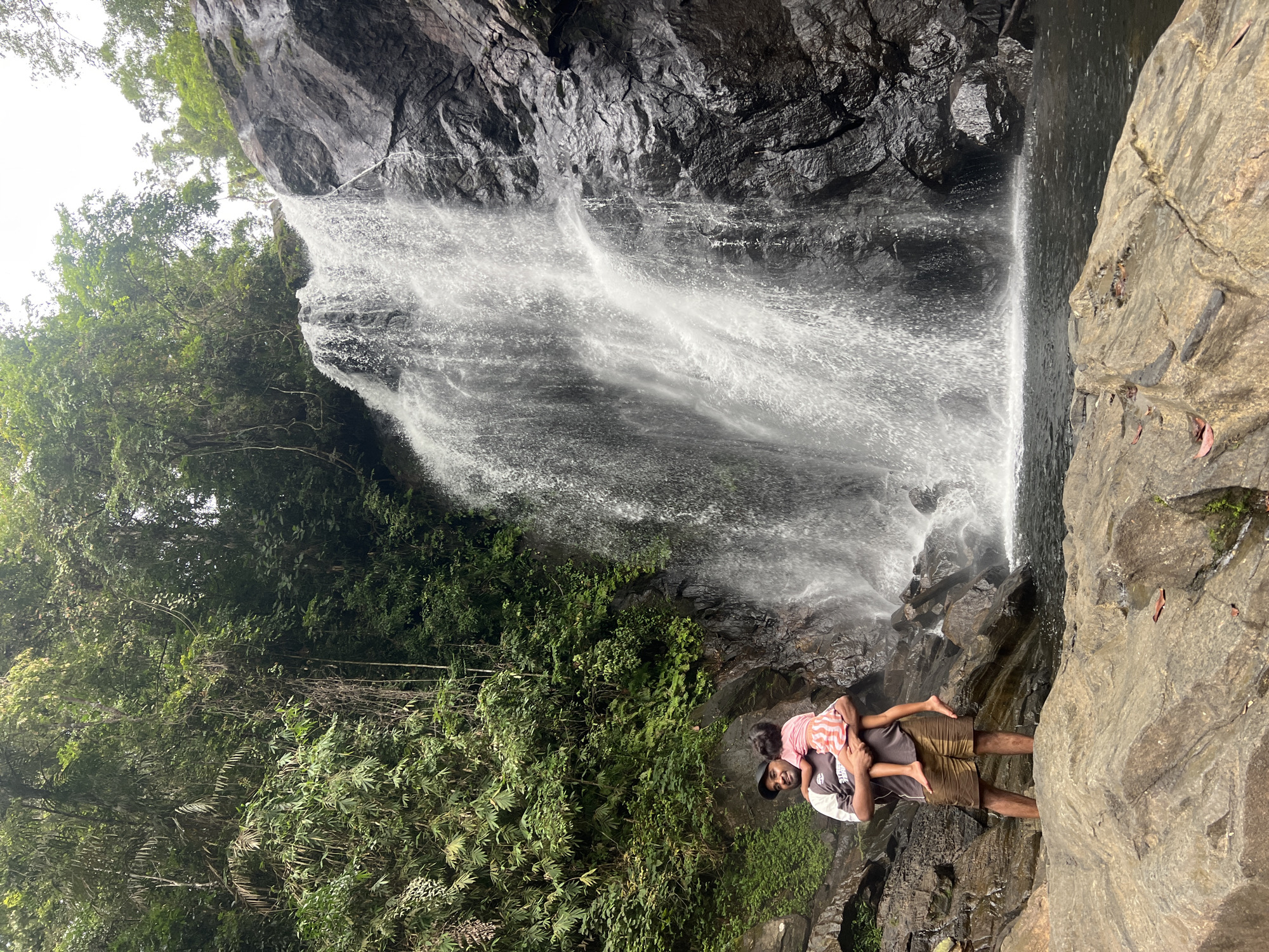 In Lankagama, Sri Lanka, a man stands in front of a cascading waterfall, holding a child. The man, wearing a dark t-shirt and shorts, is looking towards the camera with a smile. The child, dressed in a pink and white striped dress, is being held by the man, facing away from the camera. The waterfall, a prominent feature of the scene, plunges down a rocky cliff face, surrounded by lush green foliage. The area appears to be a natural, tropical environment, with rocks and water forming the foreground and the waterfall and jungle forming the background. The lighting suggests an overcast day.
