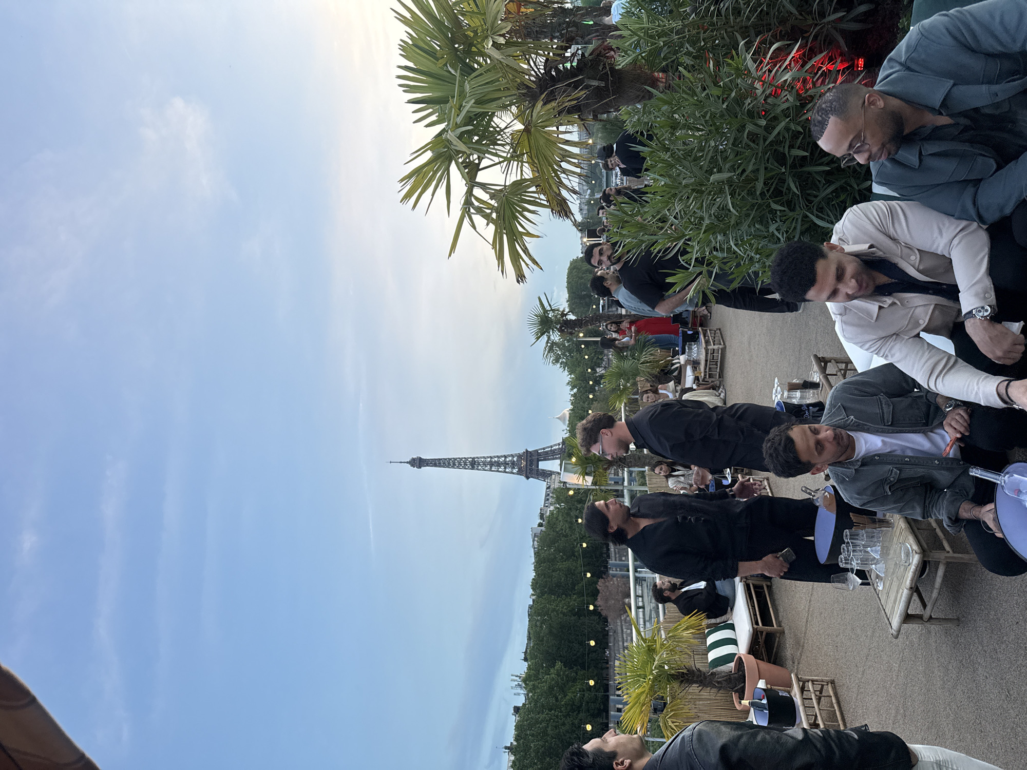 The image captures a gathering of people at an outdoor location in Paris, France, with the Eiffel Tower visible in the background. The setting appears to be a rooftop or elevated terrace with palm trees and other plants decorating the space. Several people are sitting at tables, engaging in conversations and socializing. Some are dressed in jackets and casual attire. Tables are holding drinks and glasses, indicating a possible social event or gathering. The sky is visible in the background, suggesting it may be dusk or early evening.
