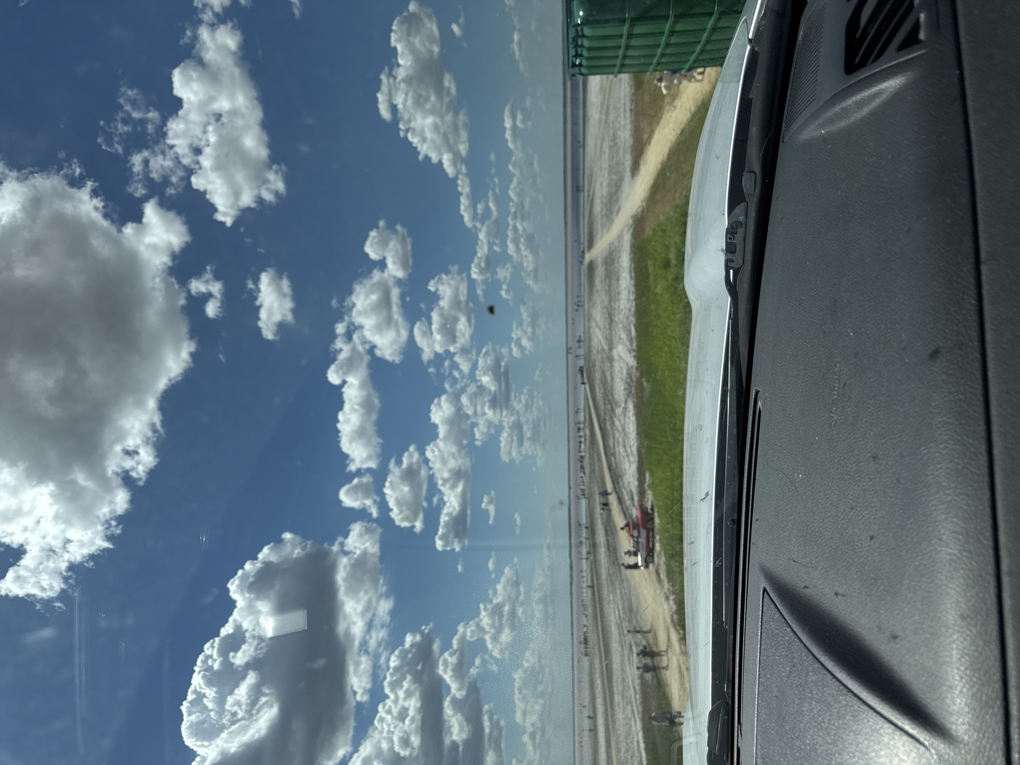 The image is a view from inside a vehicle looking out at a salt lake in Bursol, Russia. The sky is bright blue with fluffy white clouds. In the distance, a vast expanse of salt flats stretches out, dotted with numerous people. A dirt path leads towards the lake, and a red and white car is parked on the side of the path. Green vegetation lines the edge of the salt flats closest to the vehicle. A large green cylindrical object, possibly a dumpster, is visible on the right side of the image. The foreground shows the dashboard of the vehicle.