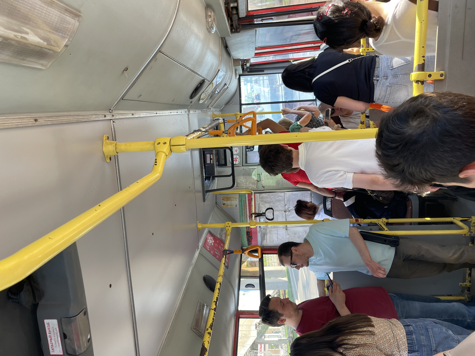 The image shows the interior of a public bus, likely in Kazan, Russia. Several people are standing, likely due to overcrowding. There are yellow handrails along the walls and ceiling for passengers to hold. A few people are looking at their phones. The environment appears to be a city street, visible through the windows.
