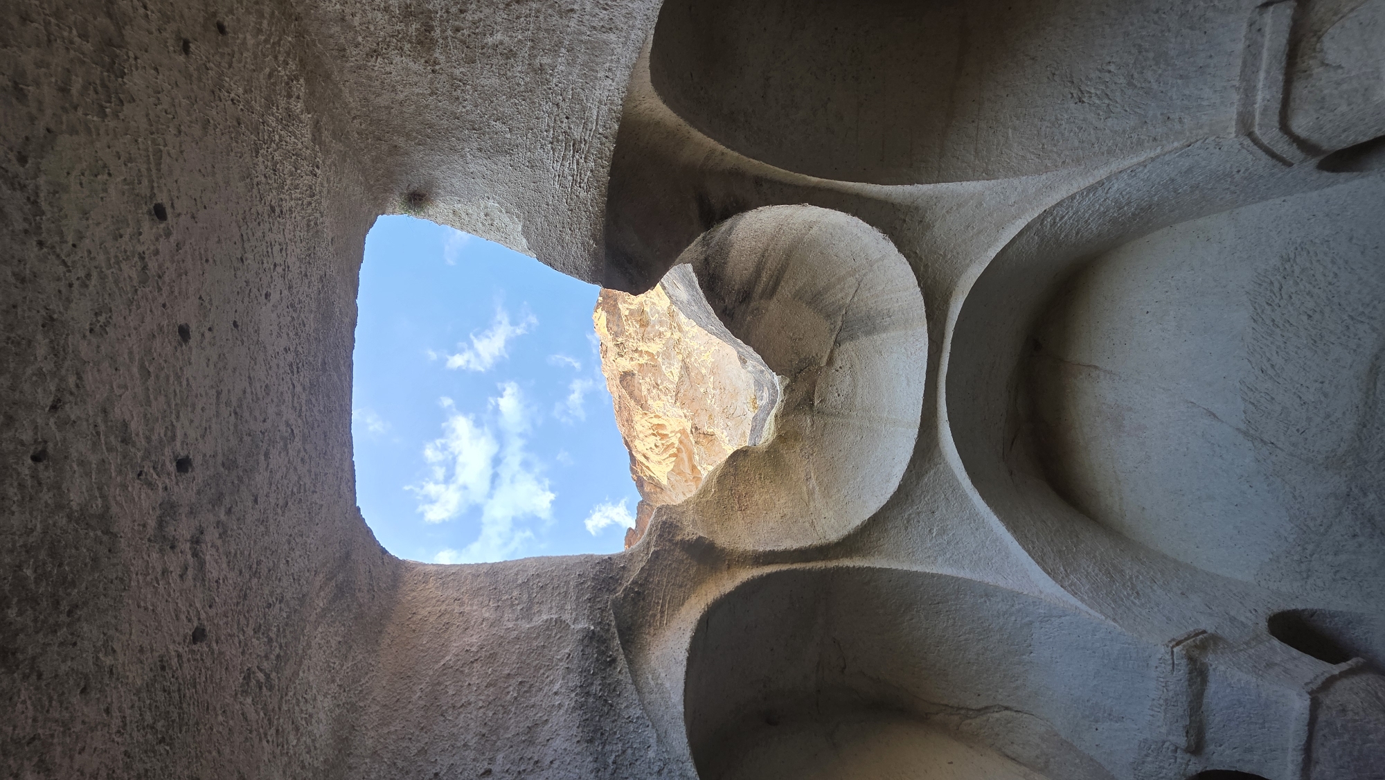The image captures an upward-looking perspective from within a carved rock structure, characteristic of Ürgüp, Turkey. The foreground features the textured, light-colored stone walls of an interior space, which exhibit both rough, natural rock surfaces and smoother, intricately carved curvilinear forms. These carvings create organic arches, recesses, and undulating patterns on the right side of the frame, suggesting a combination of natural erosion and skilled human craftsmanship.
Through a large, irregular opening towards the top-left and center, a vibrant blue sky with scattered white cumulus clouds is clearly visible, serving as a bright focal point. Adjacent to this sky view, to the right, another smaller opening reveals a sun-drenched exterior rock formation, appearing in warm golden and orange hues due to direct sunlight. This provides a stark contrast between the shaded interior and the brightly lit outside world.
No people or explicit activities are depicted; the image focuses on the architectural and natural forms. The scene is illuminated by bright daylight, indicating it is daytime, likely mid-morning or early afternoon, given the intensity and angle of the light. The weather appears clear with some cloud cover. The unique rock formations and carved structures strongly suggest a historical dwelling, monastery, or complex found in the Cappadocia region. There is no visible text in the media file.