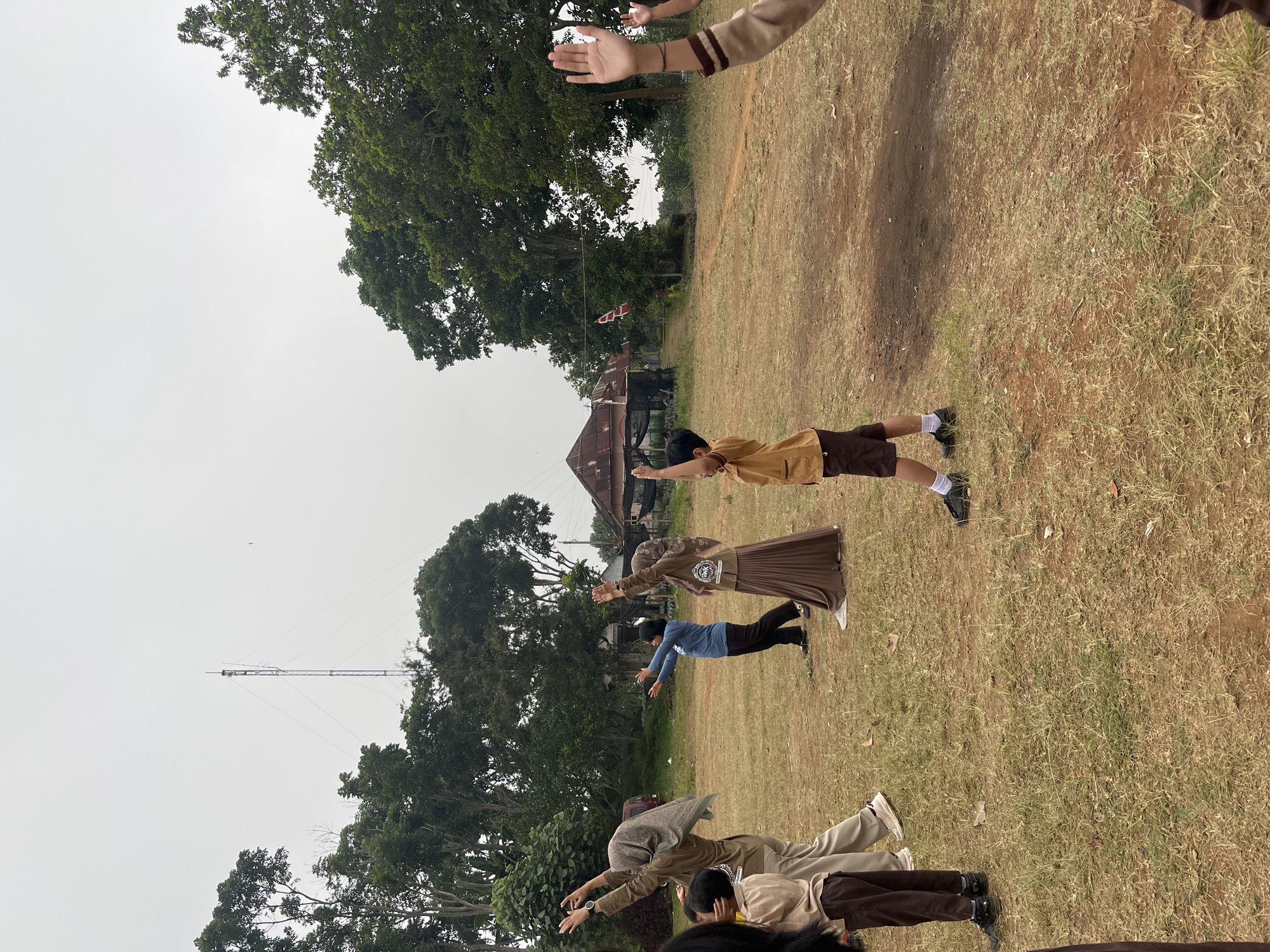 In Gembong, Indonesia, a group of individuals, mostly children in school uniforms, are engaged in outdoor physical activity on a grassy field. The scene is set against a backdrop of lush green trees and a cloudy sky. A tall telecommunications tower is visible in the distance, along with a rustic building with a corrugated tin roof. The children, in various poses, appear to be stretching or performing calisthenics under the guidance of what seems to be an instructor or teacher. Their uniforms consist of brown shirts and pants for the boys, and brown attire with headscarves for the girls. The ground is covered with dry grass and patches of soil, indicating a rural or semi-rural setting. The overall atmosphere suggests an educational or recreational activity taking place in an open-air environment.